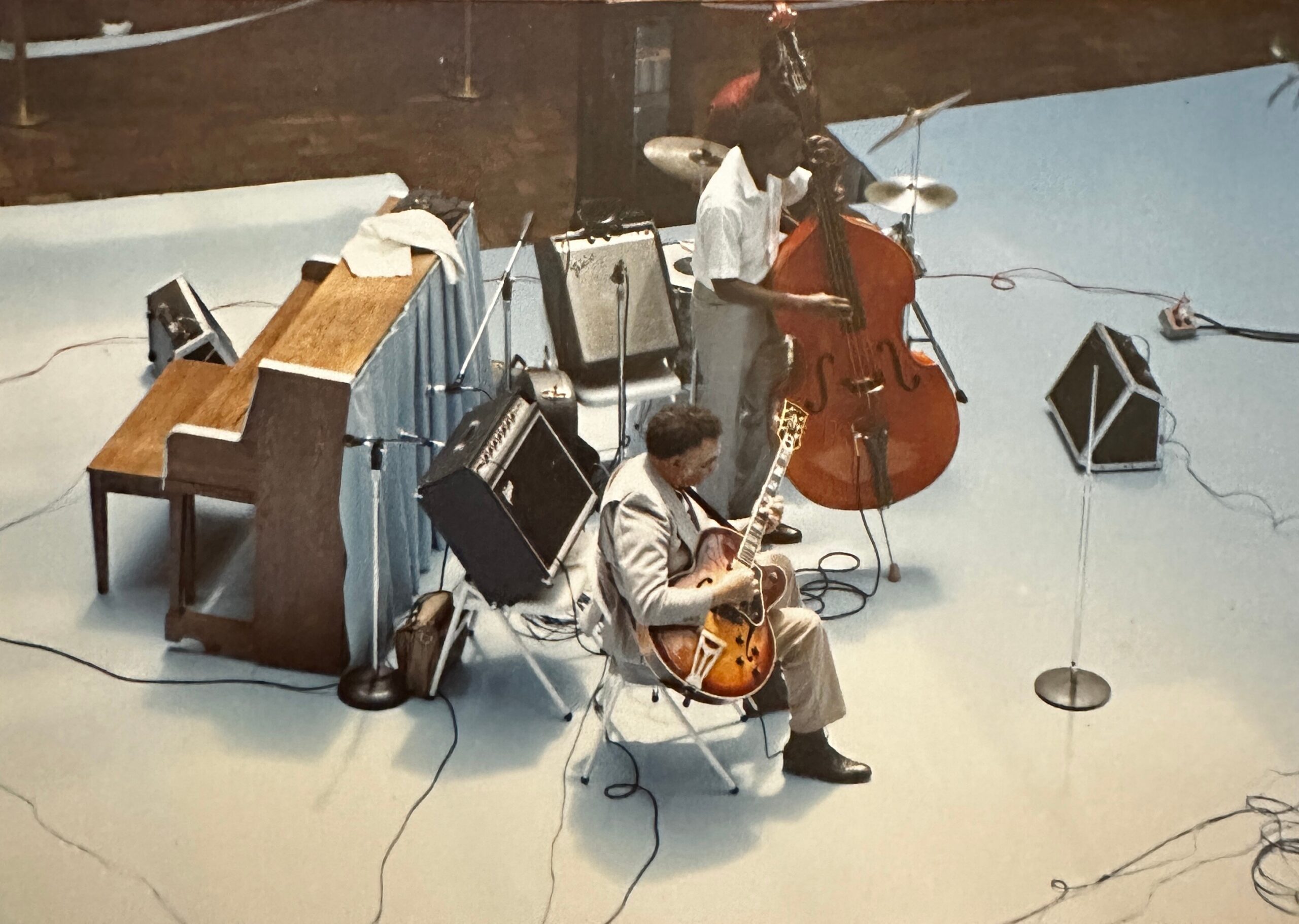 Band rehearsal in a studio: a bass player standing and others with guitars, drums, and amplifiers scattered around with cables on the floor.