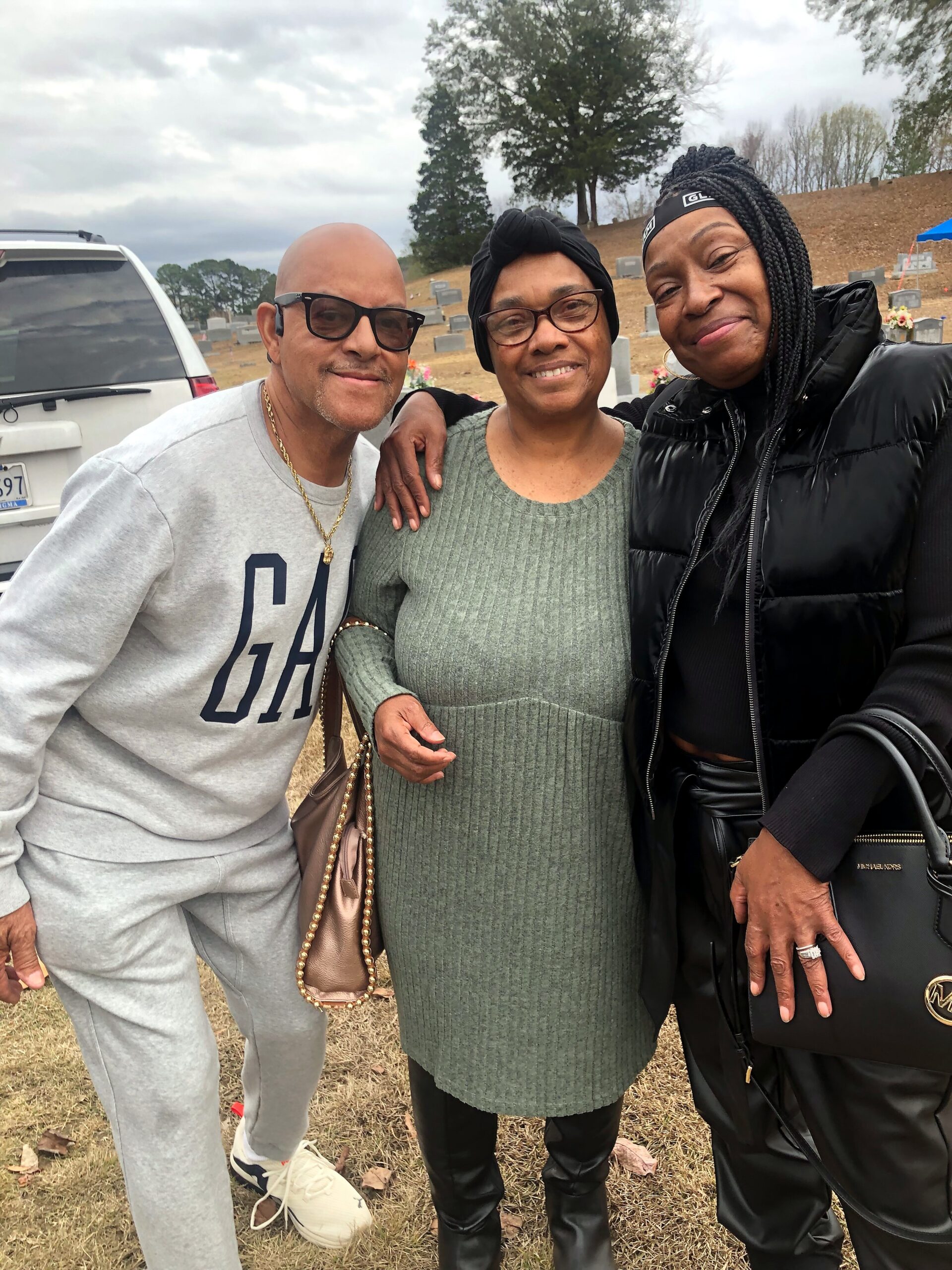Three smiling adults posing together at a cemetery with tombstones in the background. Left man in gray GA sweatshirt and glasses, center woman in green sweater, right woman in black puffer jacket.