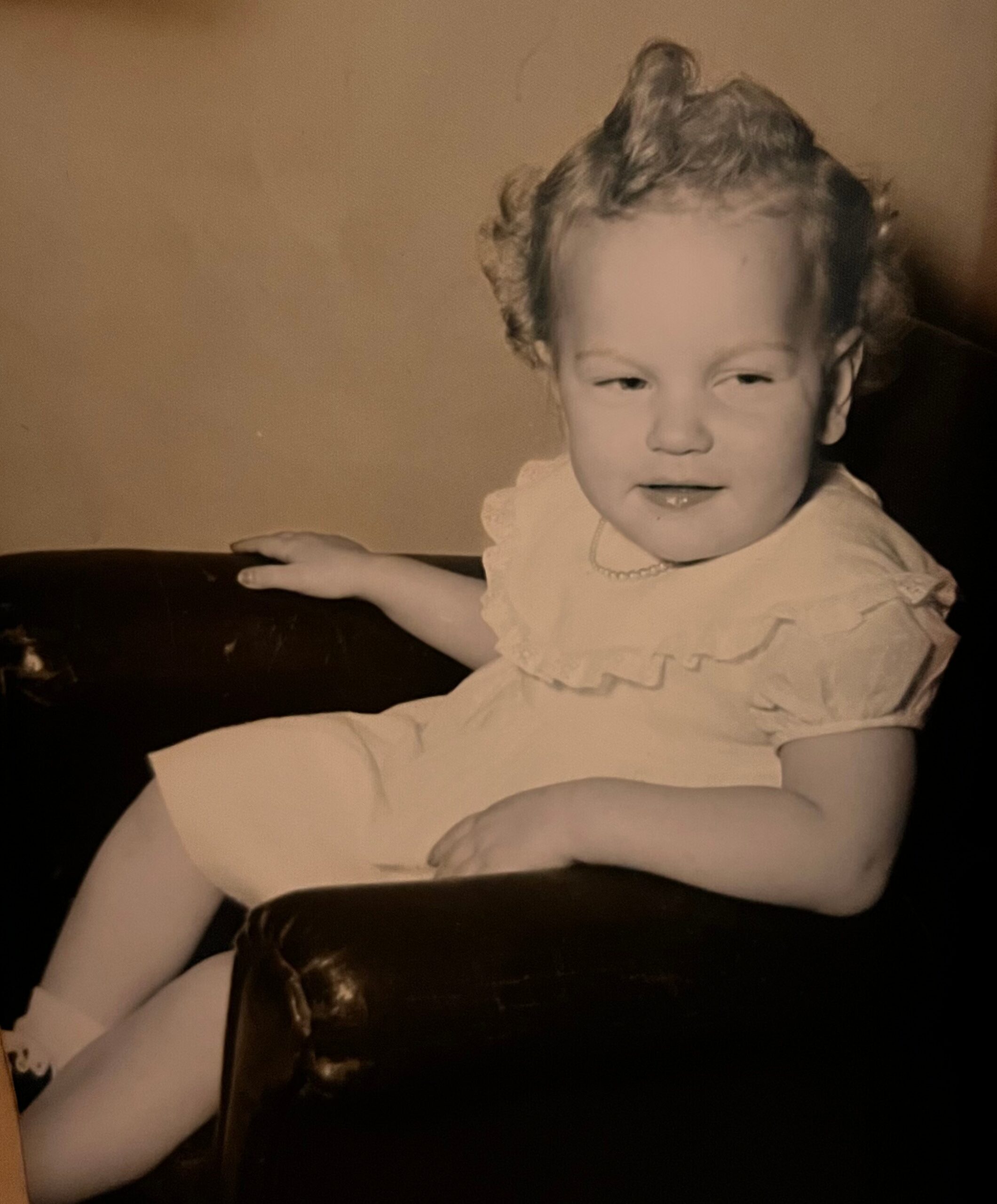 Baby sitting on a dark couch, wearing a light dress and looking toward the camera with a gentle smile.