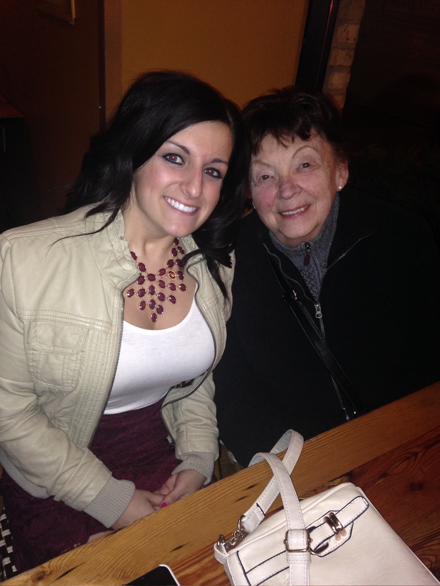 Two women smiling at a wooden table in a dim room; younger woman in a beige jacket with a burgundy necklace, older woman in a dark jacket nearby.