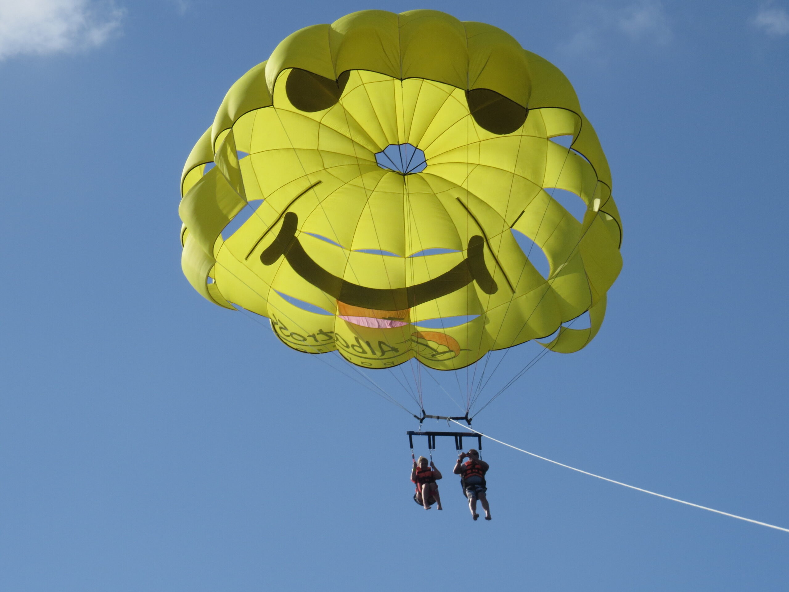 Two people parasailing under a large yellow parasail against a clear blue sky with a tow line connected to a boat below.