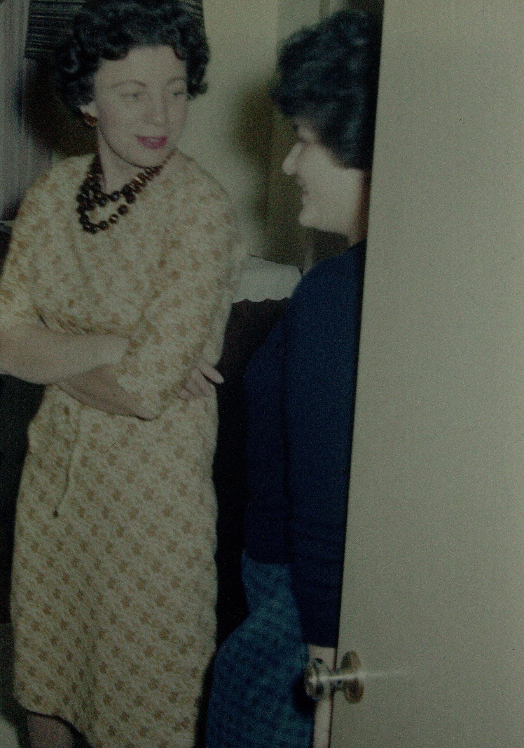 Two women in vintage outfits posing together; one in a cream lace dress with a dark beaded necklace, the other in blue, by a doorway.