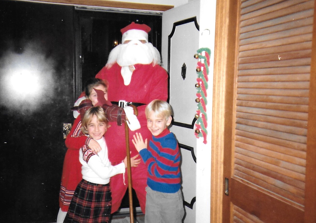 Two smiling children hug a person in a Santa suit at a doorway, with a festive garland nearby.