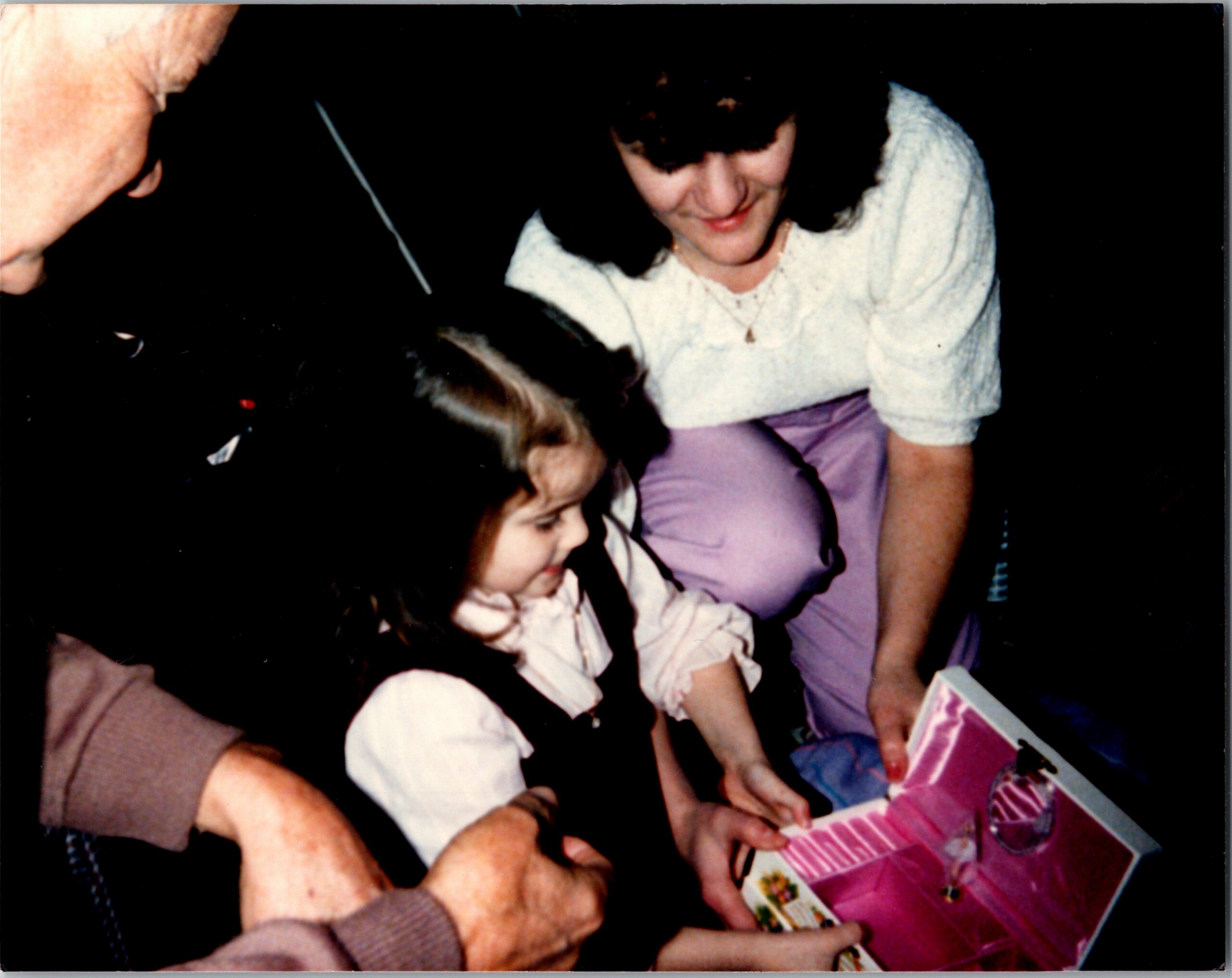 Woman with dark hair helps a young girl open a pink gift box during a celebration, smiling lightly at them.