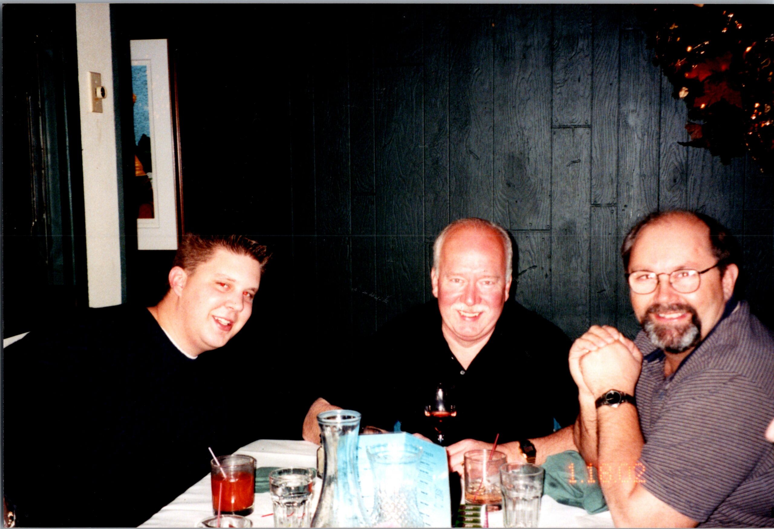 Three men sit at a table, smiling for the camera, with drinks and a blue menu on the table in a casual restaurant setting, likely friends.