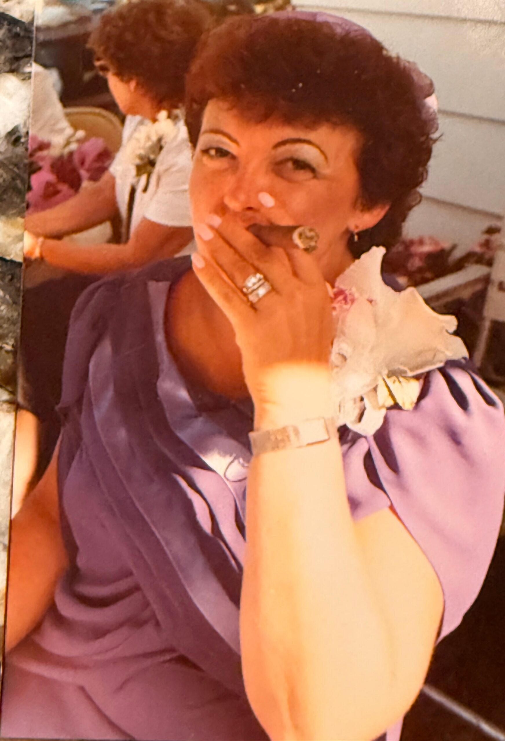 Woman with short dark hair in a purple blouse covering her mouth with her hand, wearing rings and a watch at a social gathering.