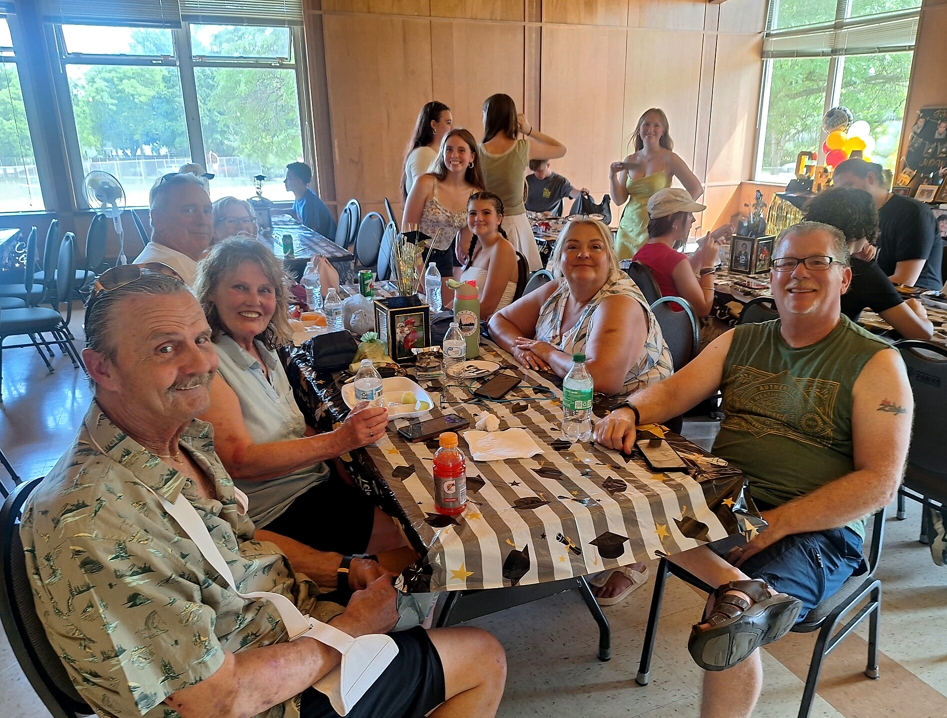 Group of people sitting at a long party table indoors, smiling for the camera with drinks and snacks on the table.