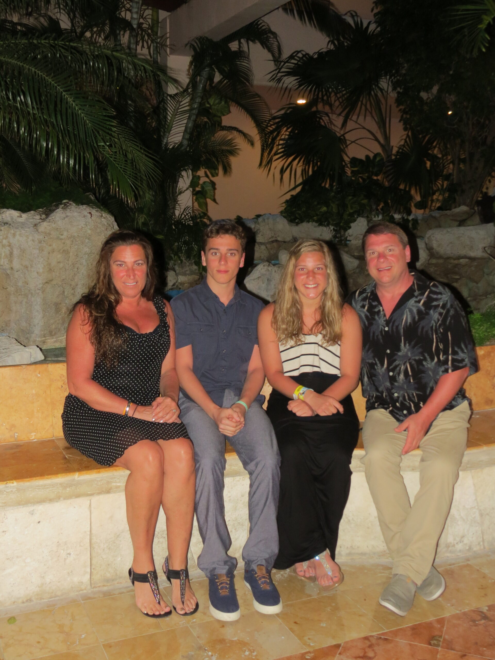 Four people sit on a stone ledge by tropical plants at night, smiling for the photo.
