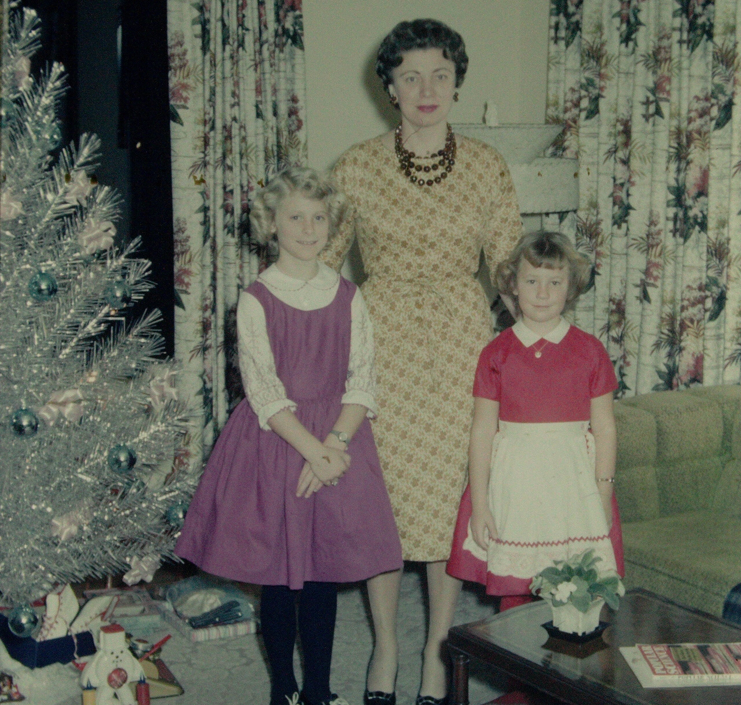 Mother in a gold patterned dress with pearl necklace posing with two children beside a decorated Christmas tree in a floral-curtained living room are captured in a vintage family photo.