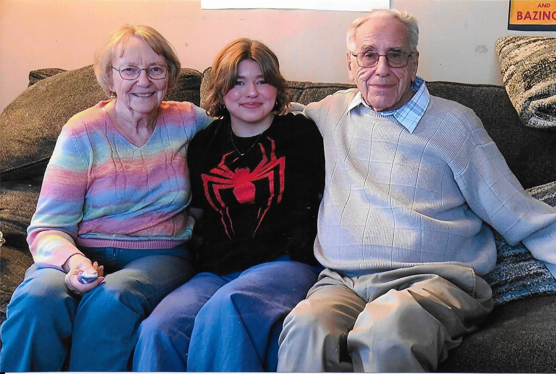Three people sit close together on a sofa smiling at the camera: a grandmother in a pastel rainbow sweater, a young woman in a Spider-Man shirt, and a grandfather in a light sweater.
