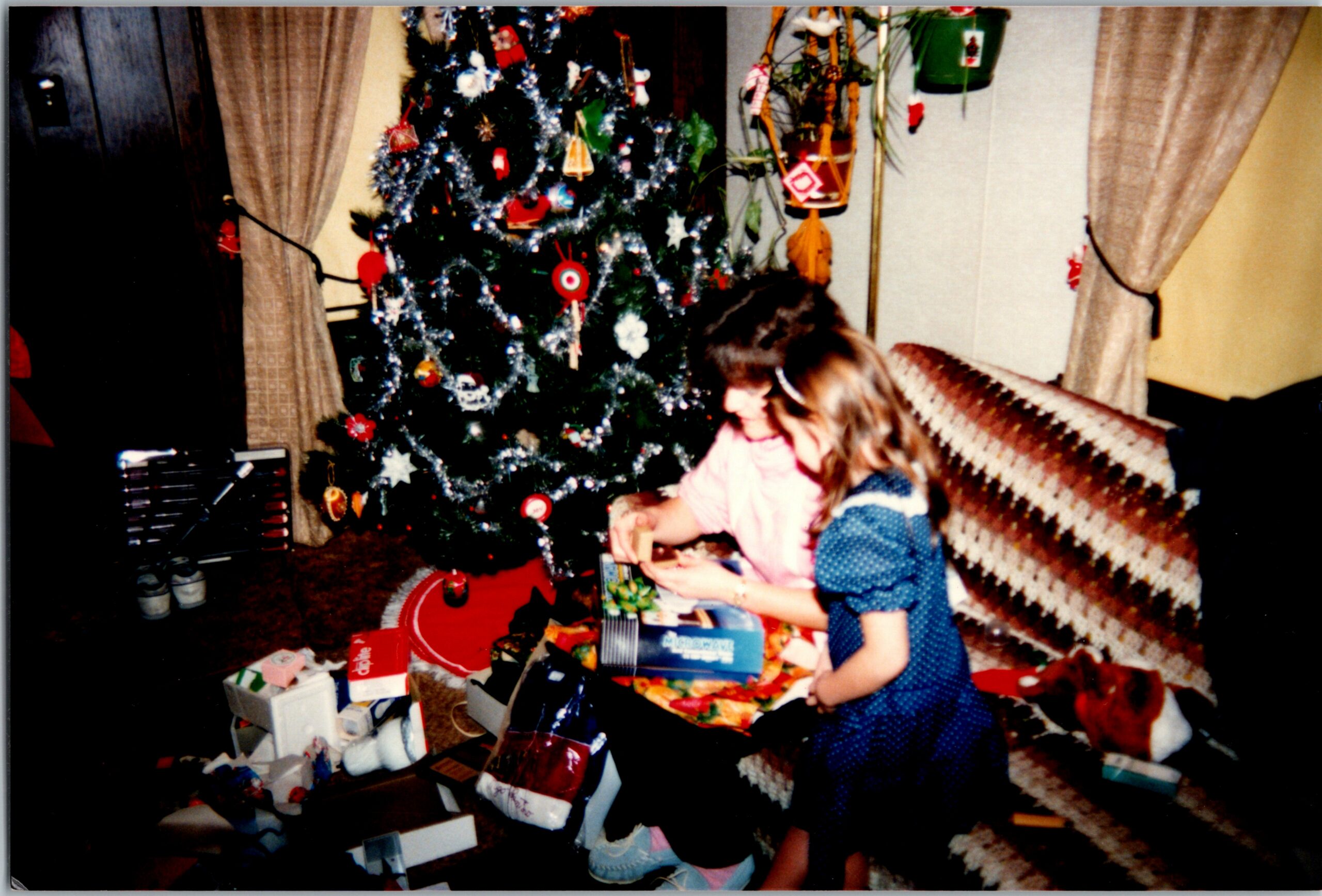 Two children sitting on the floor by a decorated Christmas tree, opening presents amid scattered gifts and wrapping paper in a cozy living room.