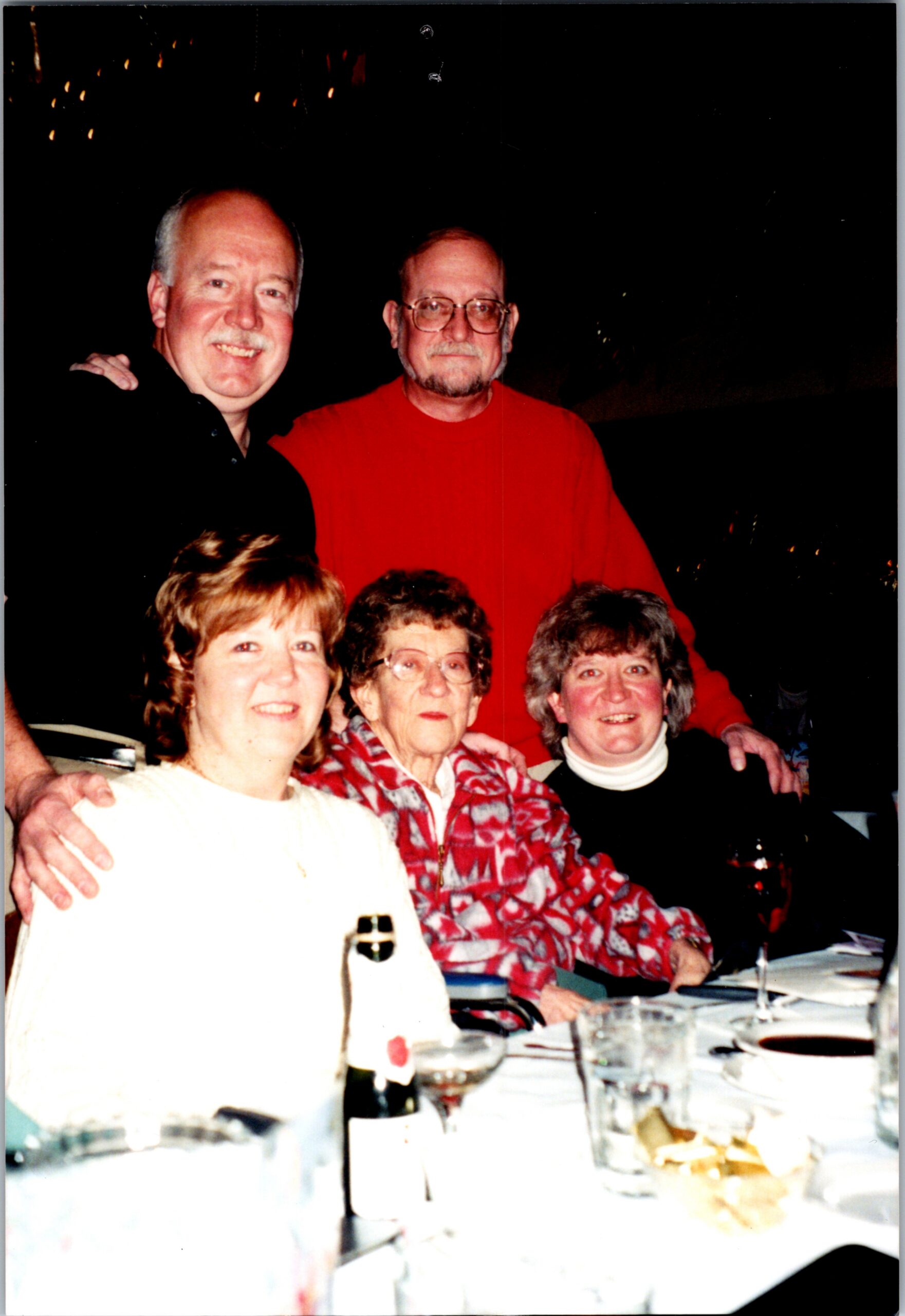Five adults pose for a dinner photo around a table; a man in a bright red shirt stands behind the others, smiling.