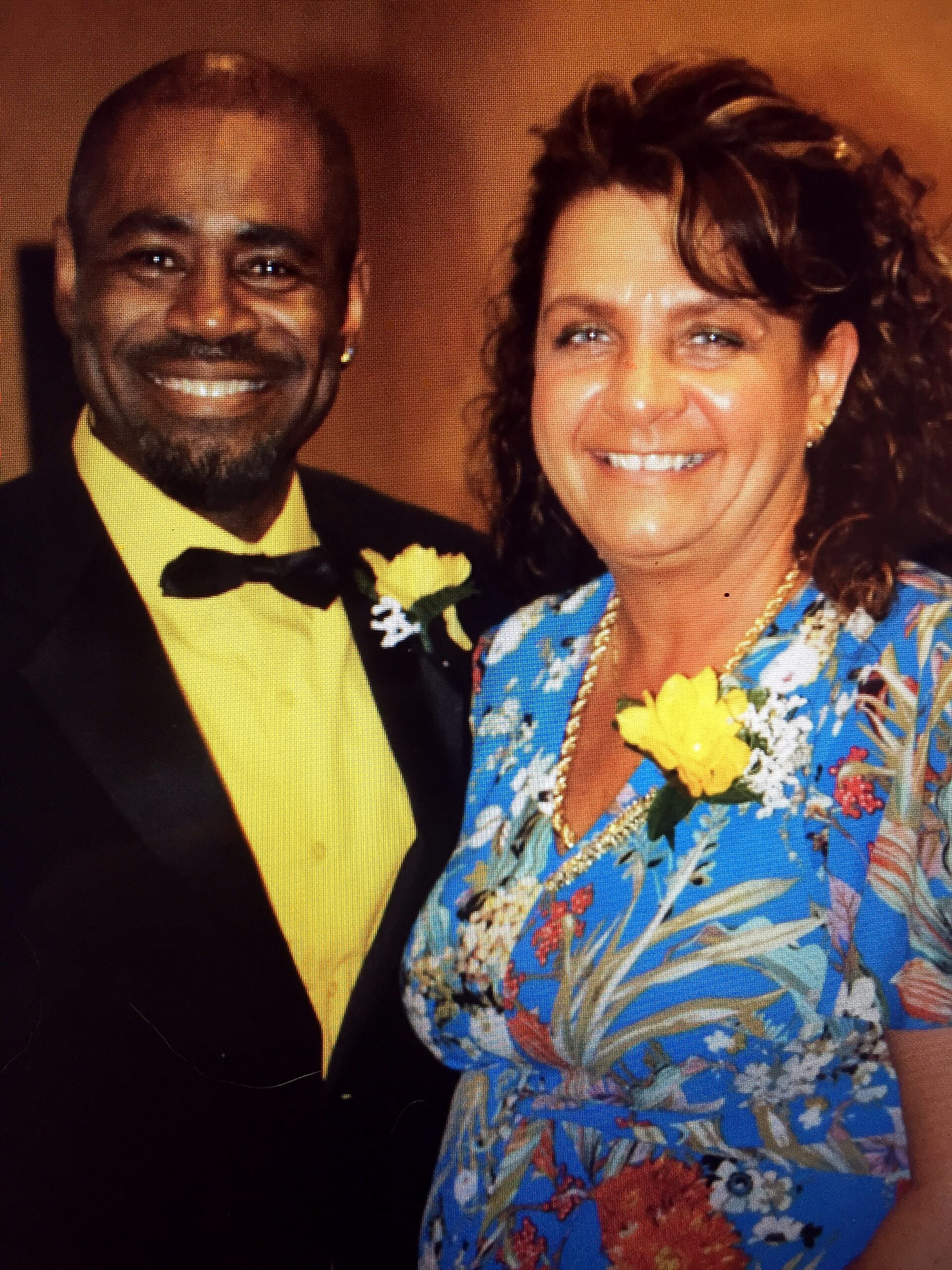 Smiling couple posing together at a formal event; man in a black suit with a yellow shirt and boutonniere, woman in a bright blue floral dress with a yellow flower bouquet visible.
