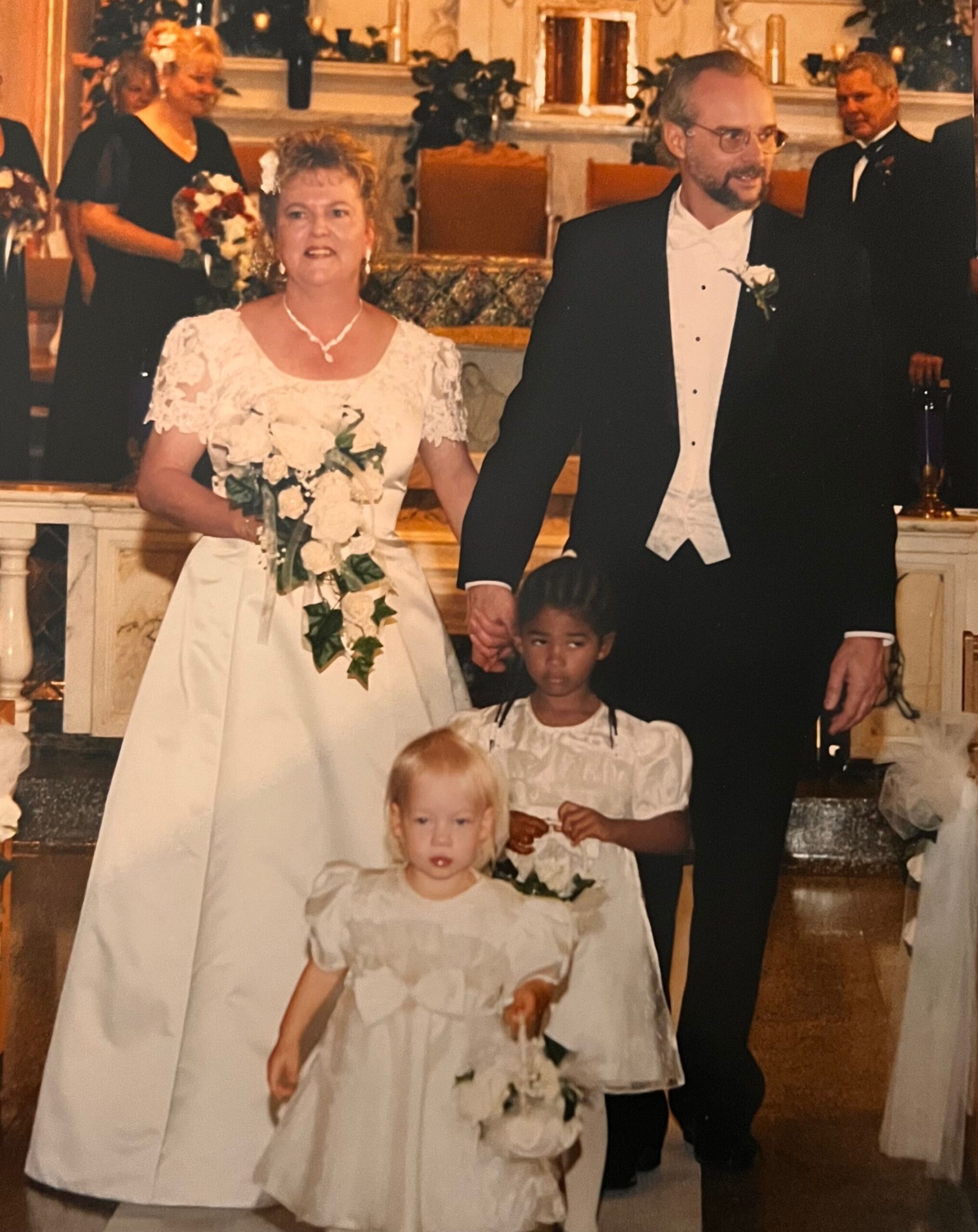 Bride in a white wedding gown holding a cascading white bouquet stands beside a man in a black tuxedo; two young girls in white dresses sit in front in a church setting.