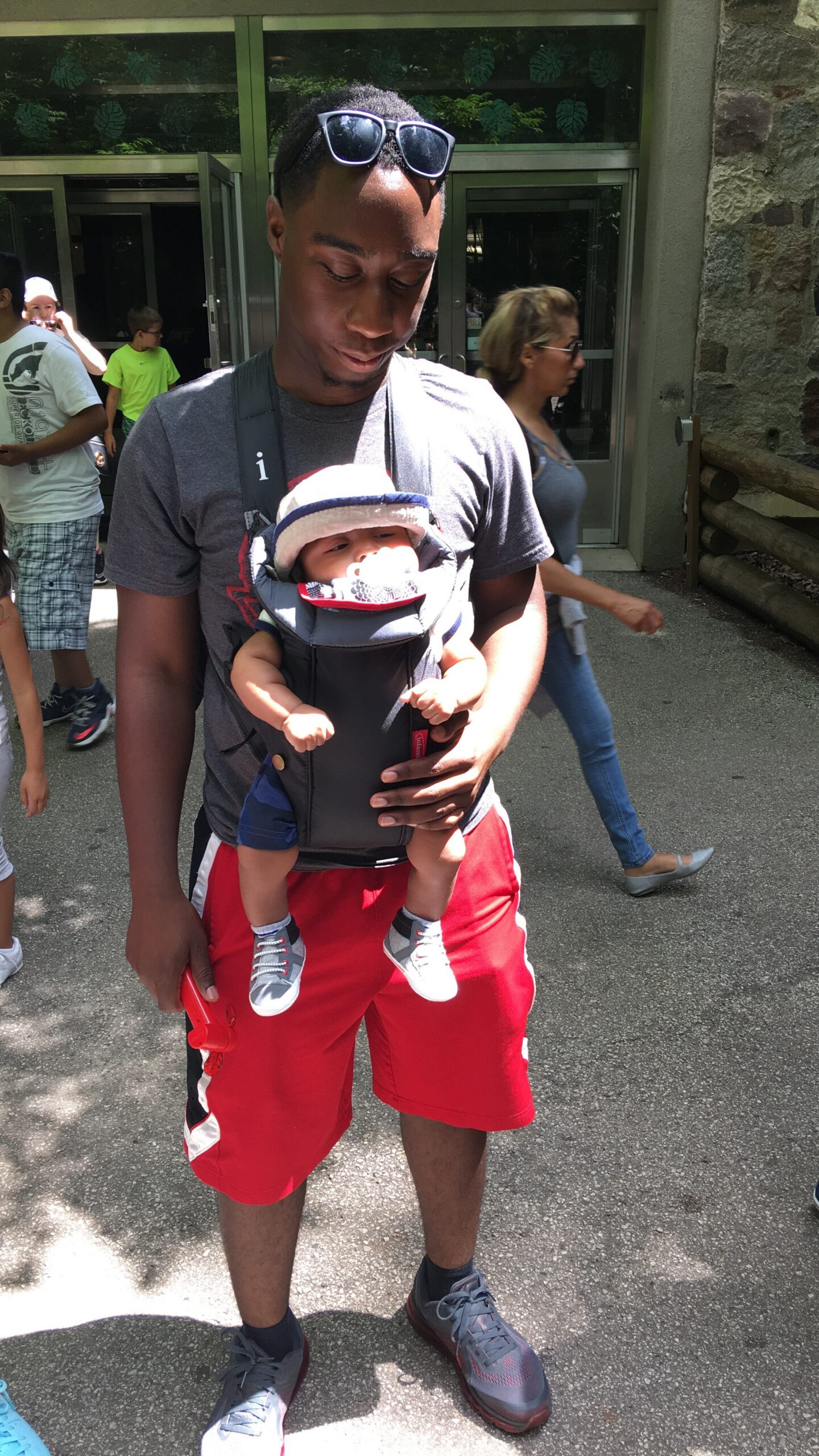 Man wearing a gray t-shirt and red shorts holds a baby in a front baby carrier outdoors, with sunglasses on his head nearby.
