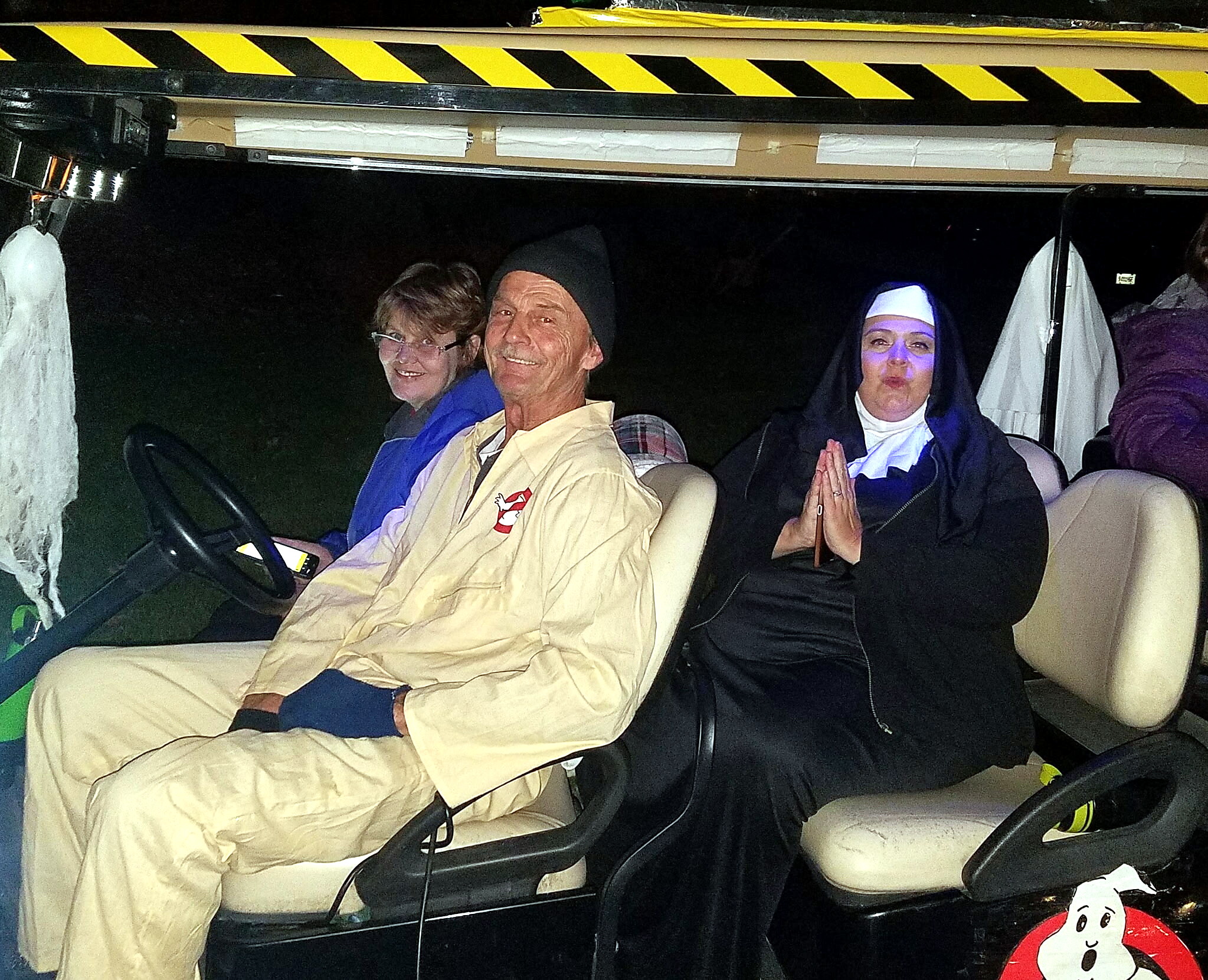 Three people in a car at night wearing costumes: a man in a beige Ghostbusters uniform, a woman in blue, and another in a nun’s outfit with a white headpiece.