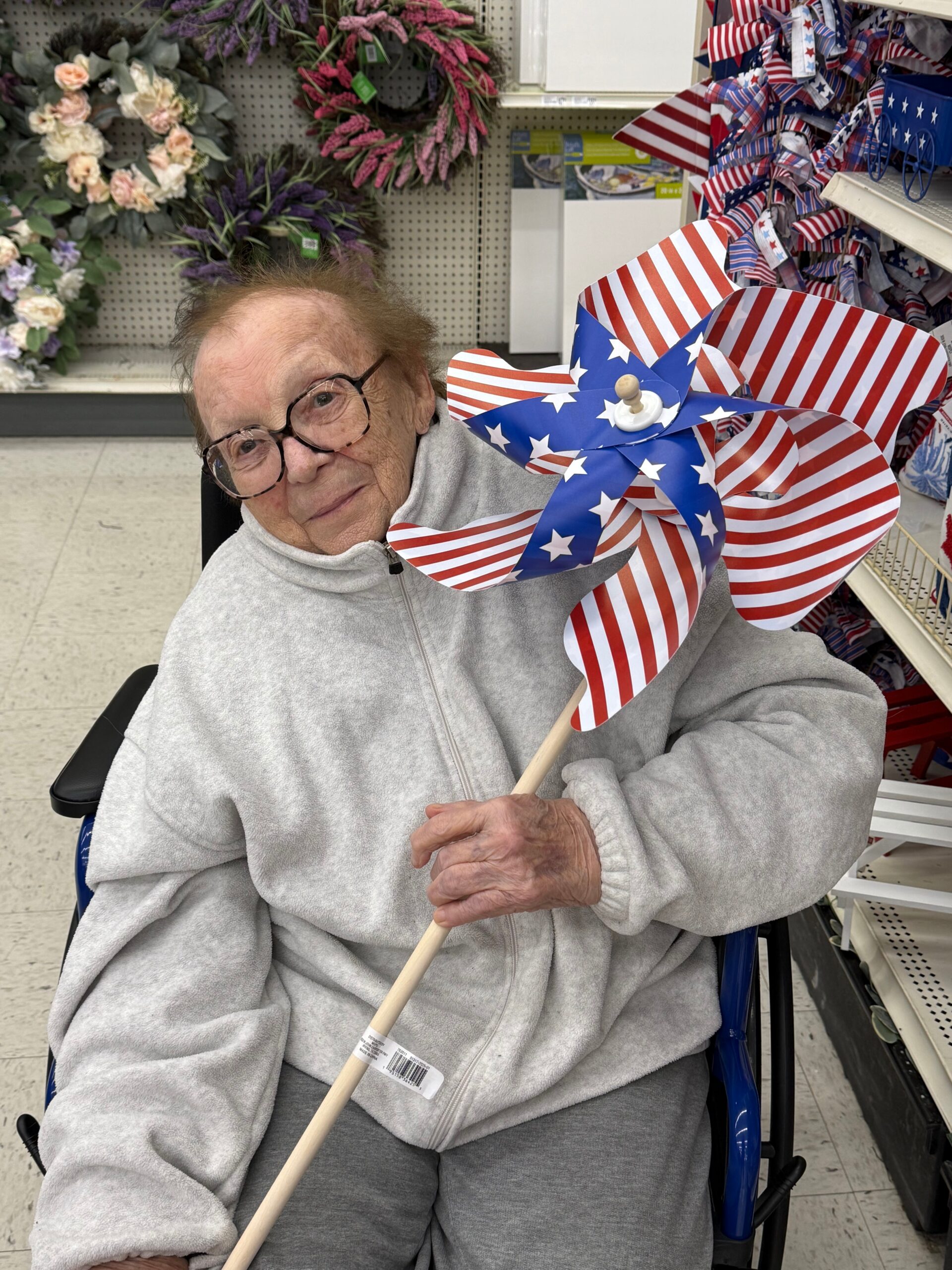 Smiling elderly woman in a gray fleece jacket sits in a wheelchair, holding a red, white, and blue star-pattern pinwheel in a store aisle.