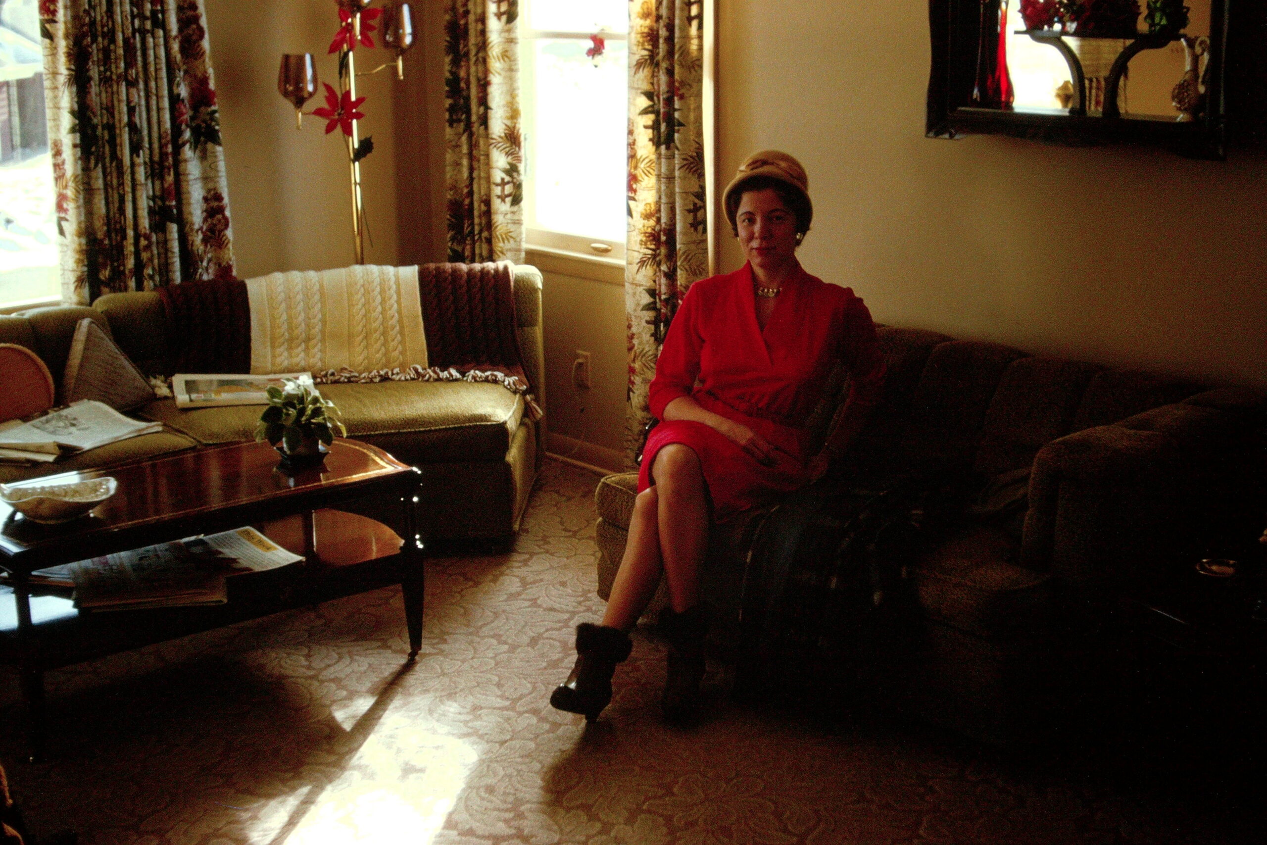 Woman in a red dress and hat sits on a sofa in a cozy living room, with patterned curtains and a coffee table nearby.
