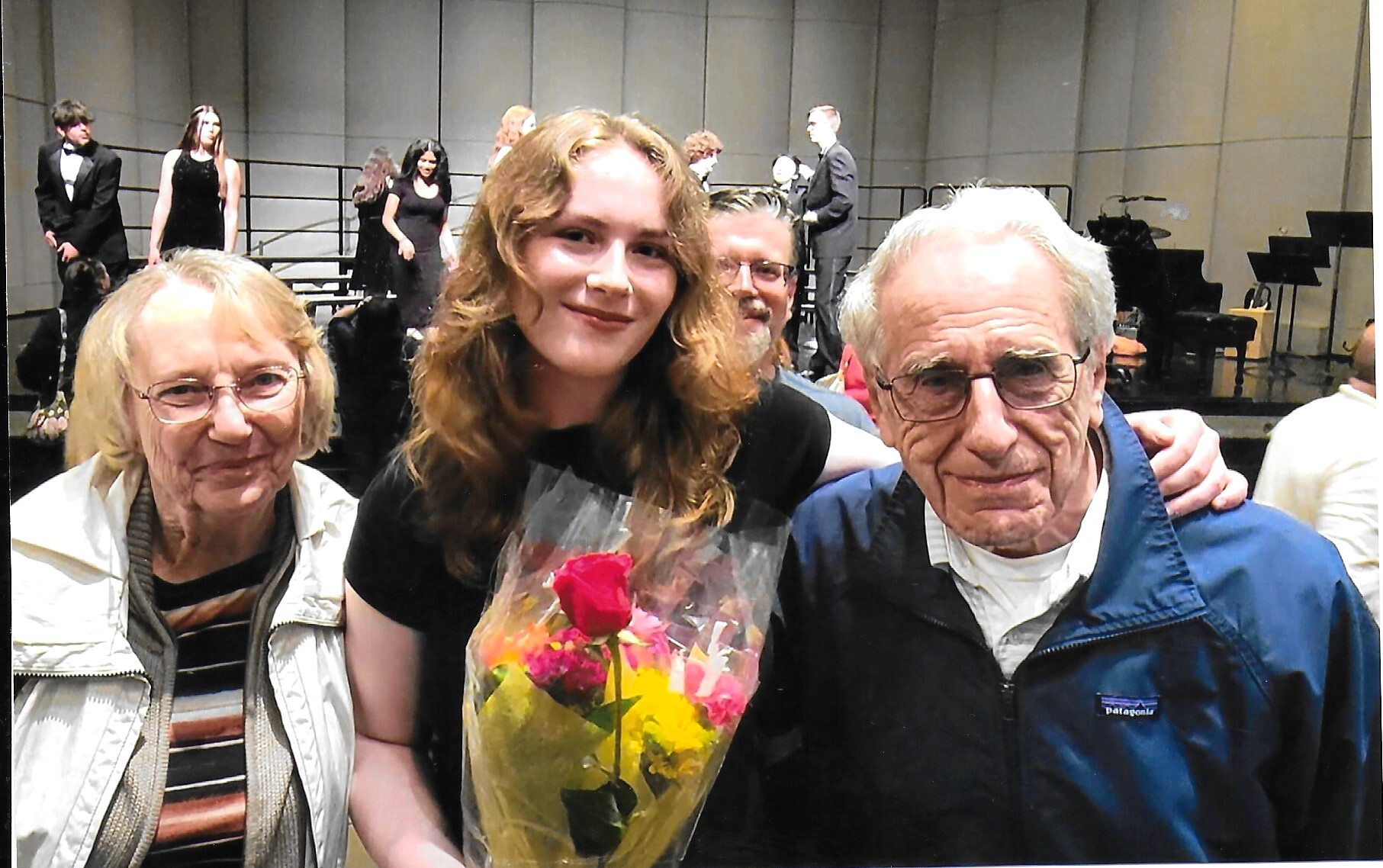 Three people pose for a photo at a recital hall; the center woman holds a bouquet while performers stand on stage in the background.
