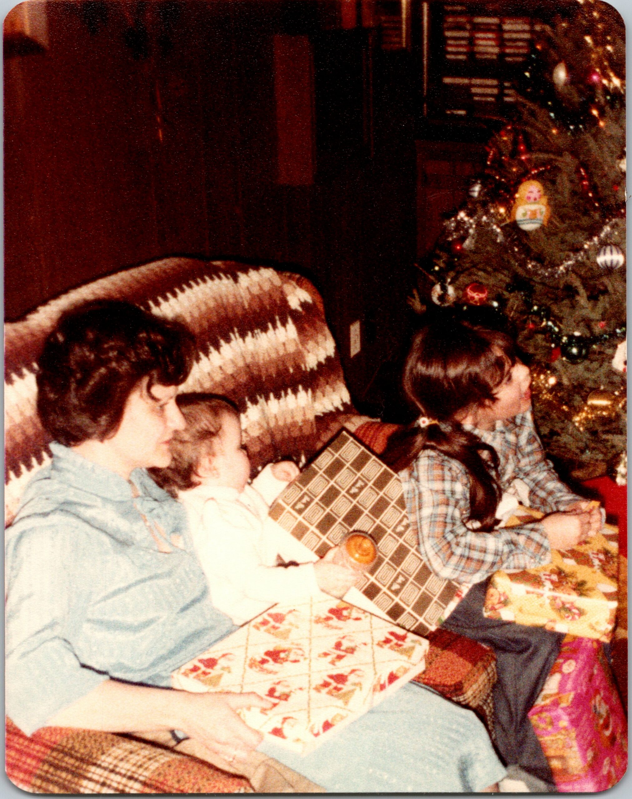 Family members sit together on a couch and floor, opening wrapped Christmas gifts near a decorated tree in a cozy living room.