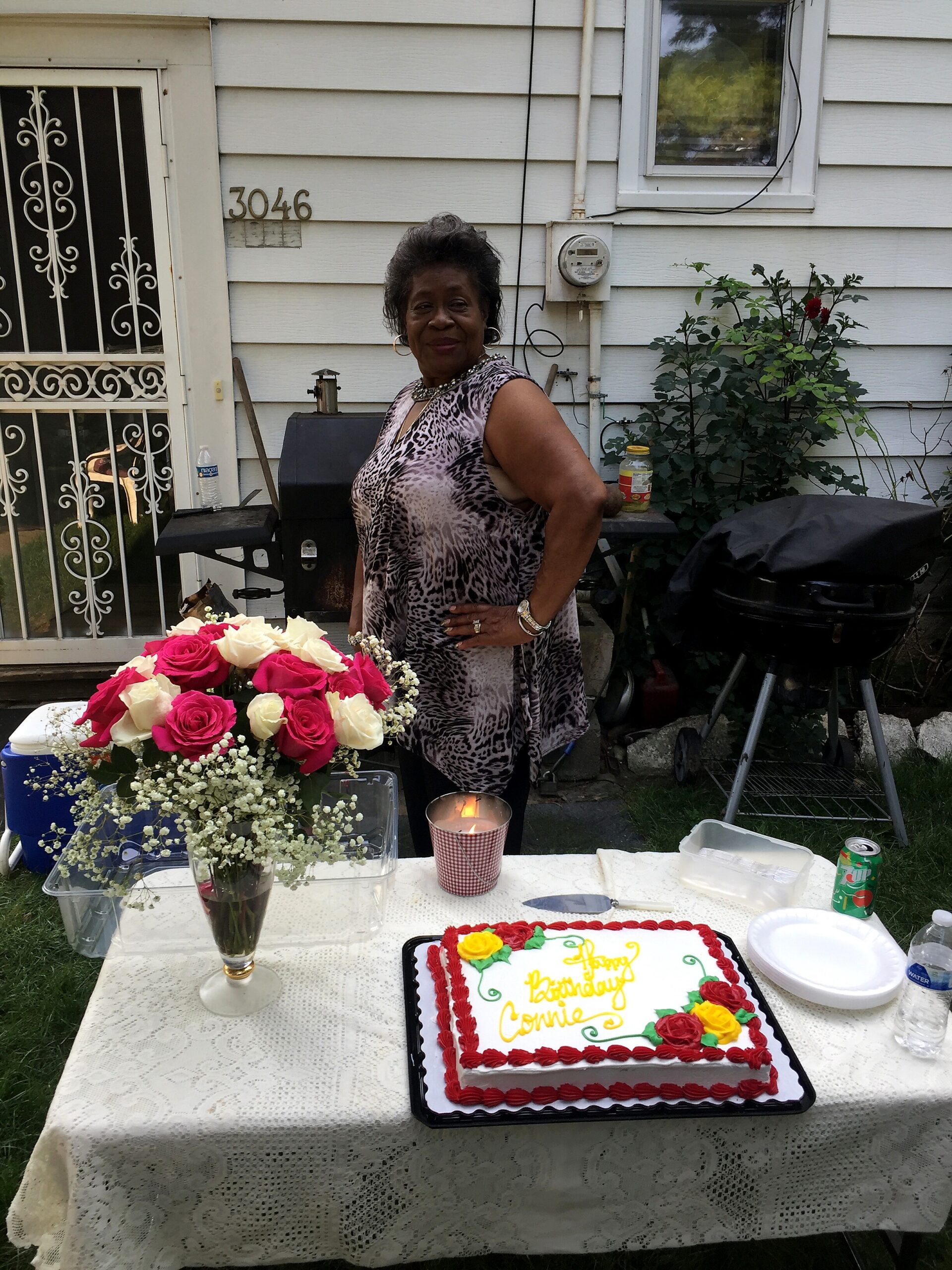 Smiling woman in a leopard-print top stands beside a table with a pink-and-white cake and pink/white flowers in a backyard birthday scene. A birthday gathering in a yard: cake on a lace tablecloth, bouquet vase, and a woman posing near a grill and house backdrop.