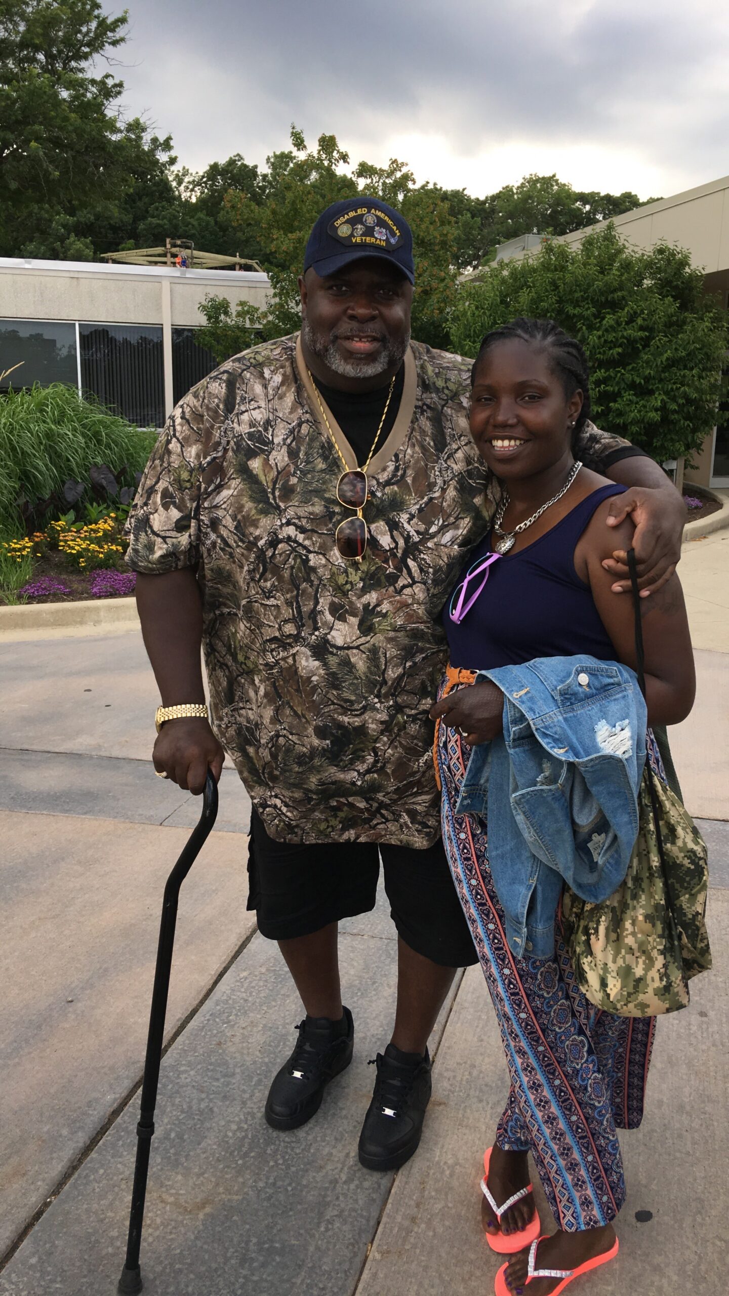 Smiling man in camouflage shirt and cap stands with woman outside; he uses a cane and sunglasses hang from his shirt.