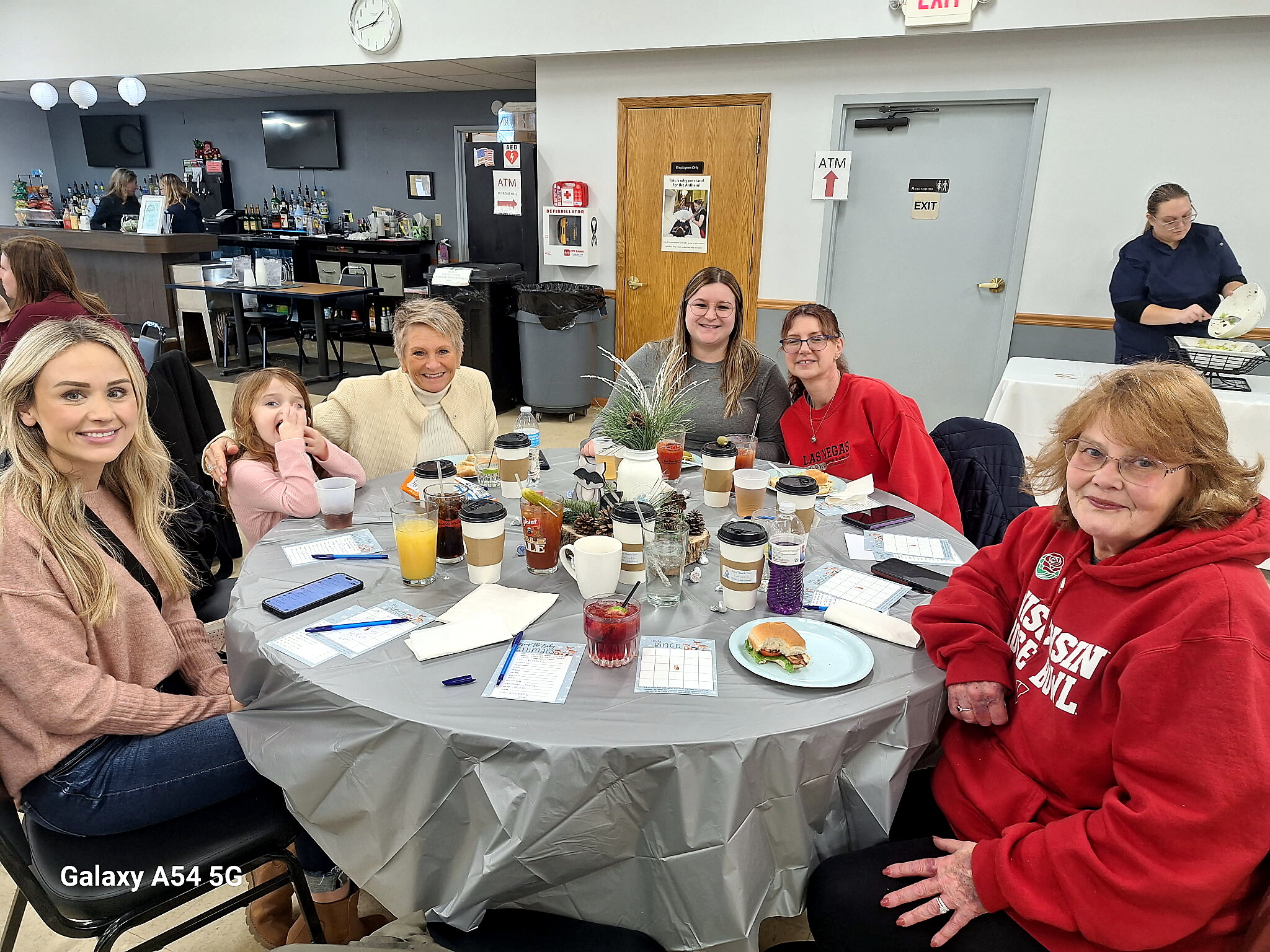 Group of people seated around a gray plastic-covered table with coffee cups, glasses, and plates during a casual meal or gathering in a hall.
