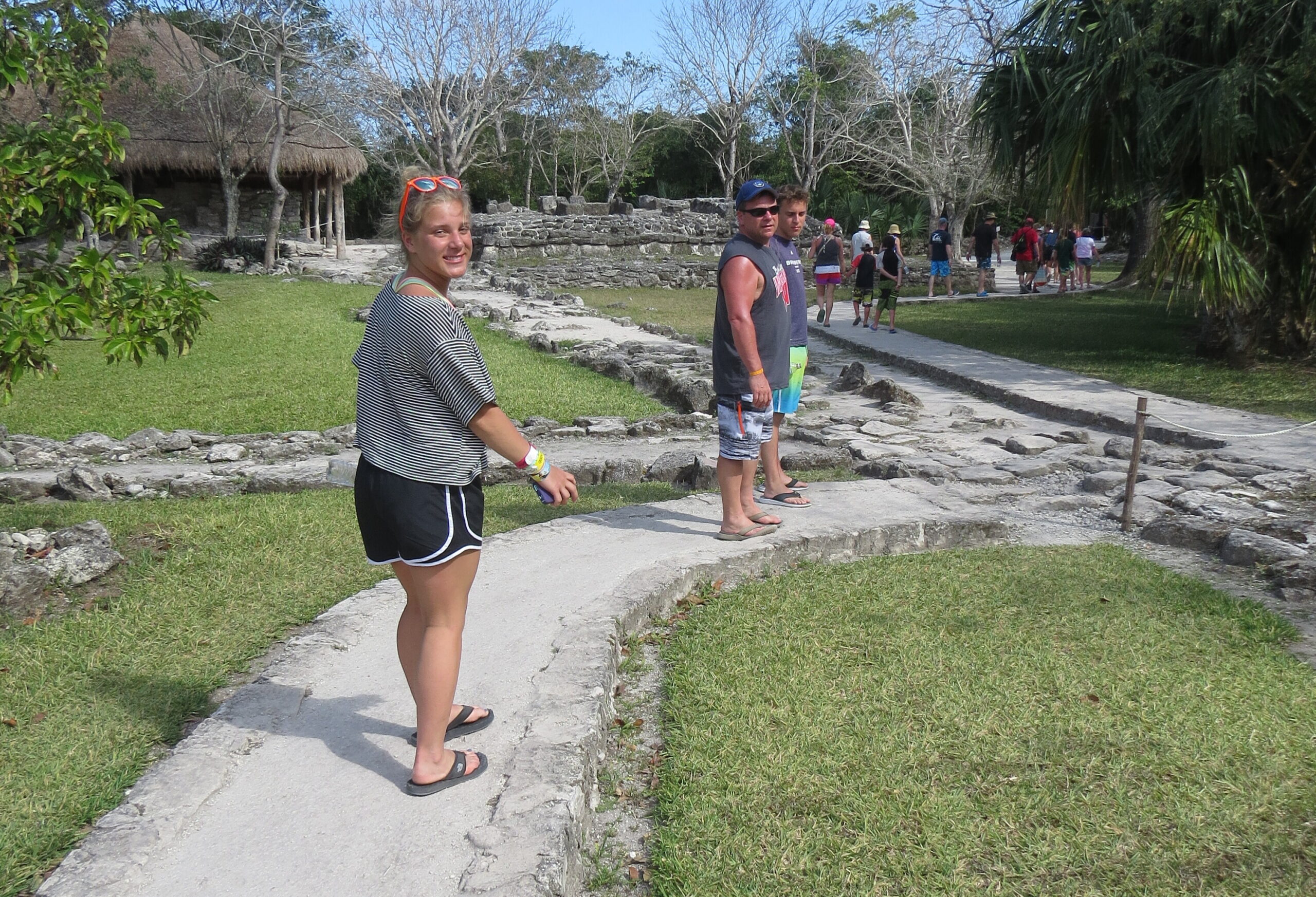 Group of tourists on a stone path near ancient ruins and palm trees; woman in striped shirt in foreground smiles at the camera.