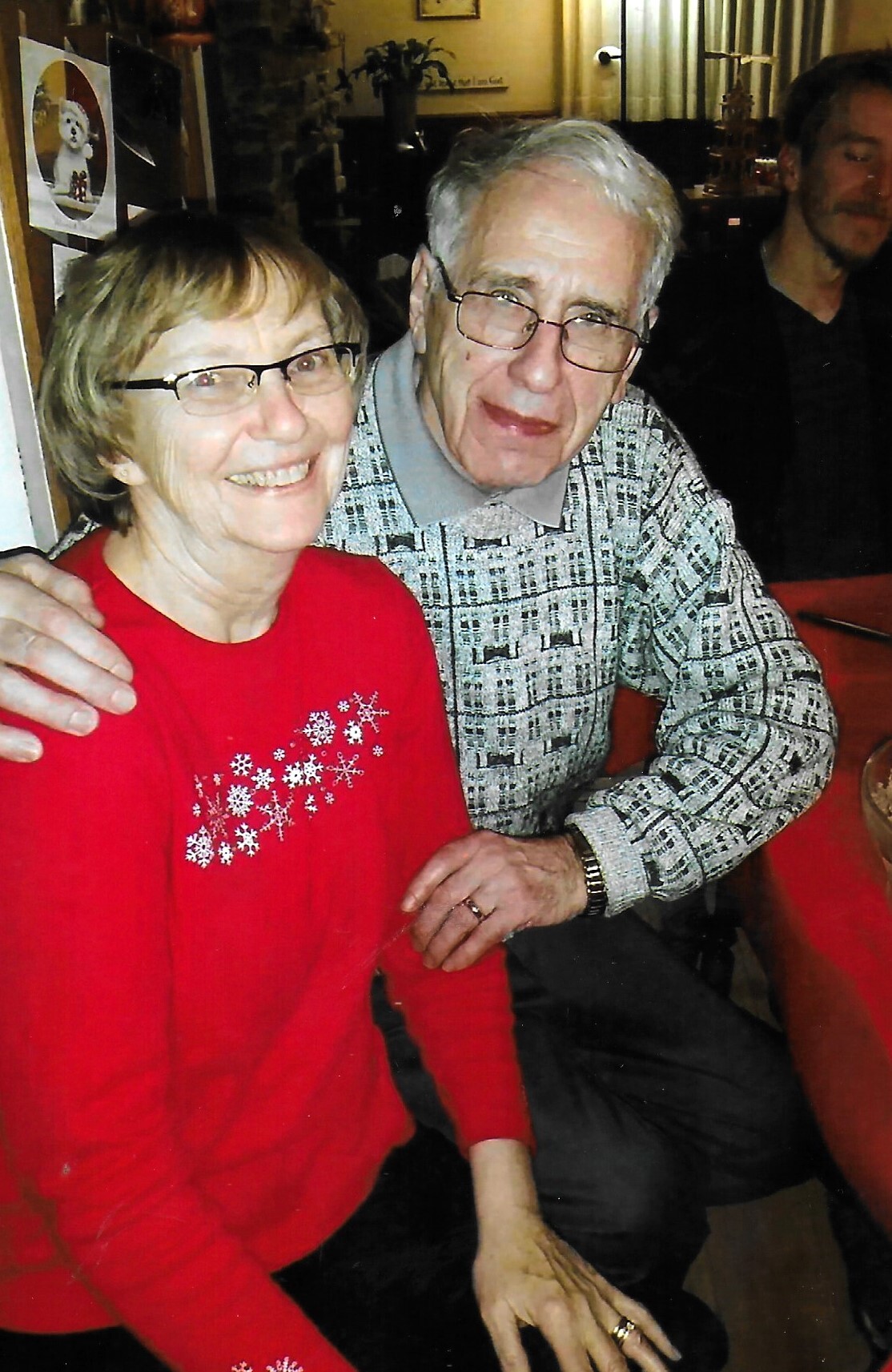 Smiling elderly couple seated closely at a dining table, woman in a red sweater with snowflake design and glasses, man in a patterned gray sweater with glasses beside her.