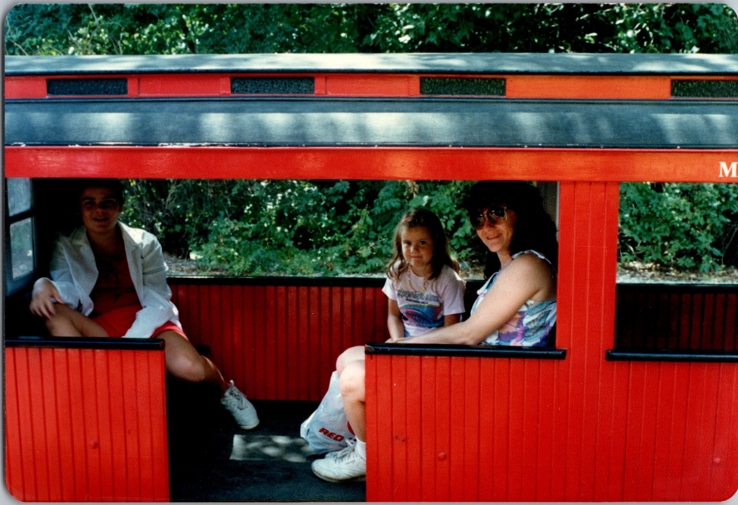 Three people seated in a red open-top tram car with greenery in the background.