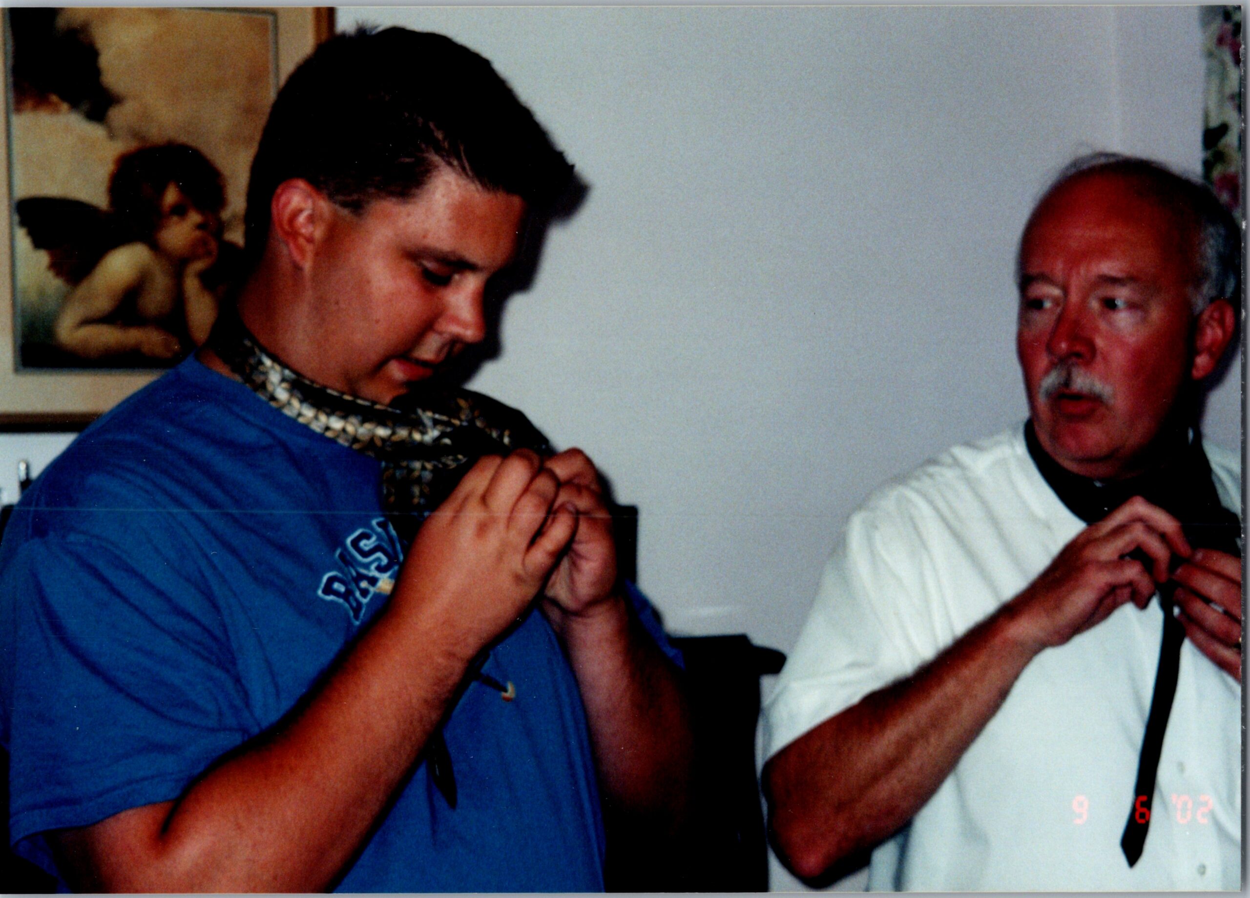 Two men clasp their hands in prayer at a casual gathering; one in a blue shirt with a chunky chain, the other in a white shirt.