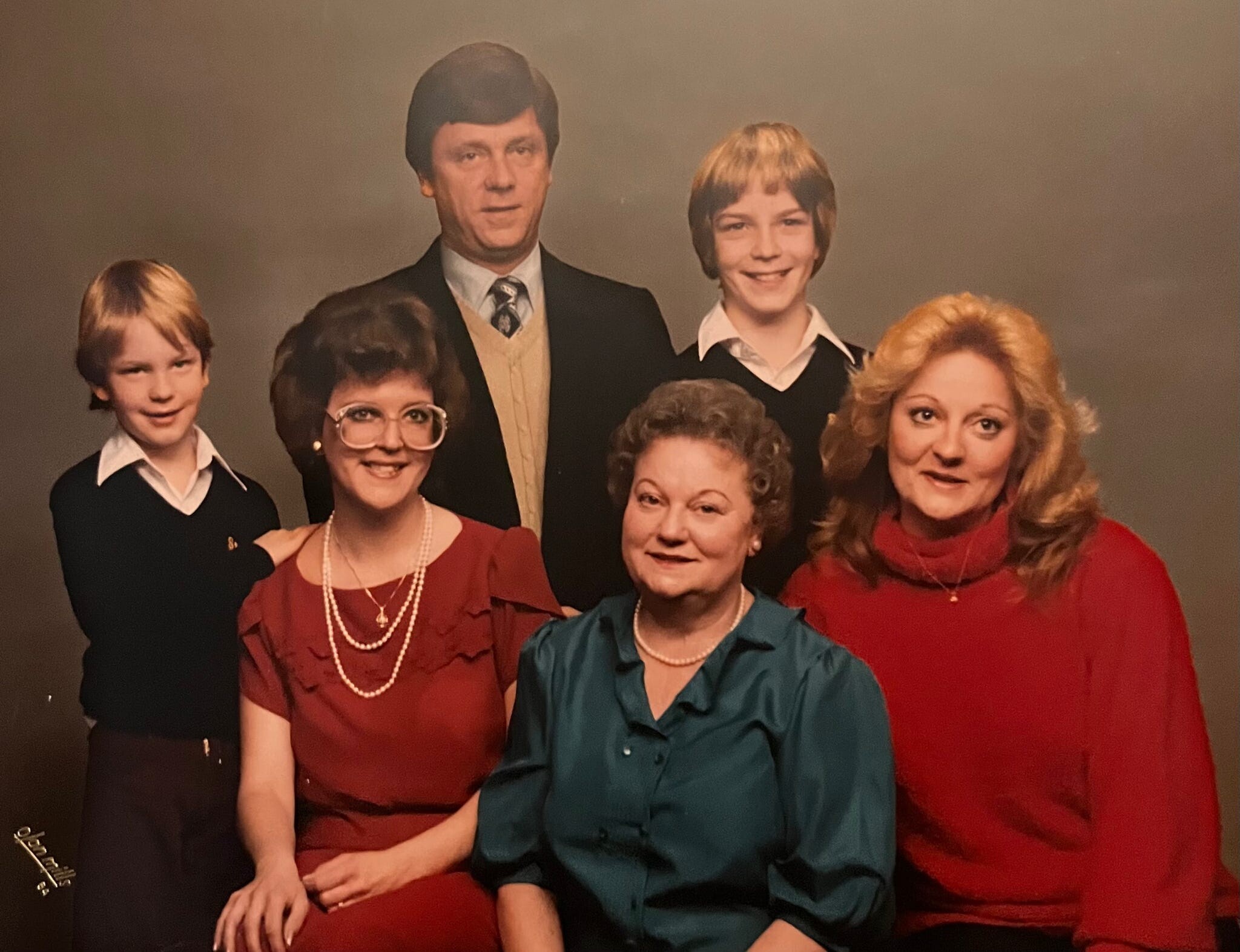 Family portrait of six people in formal attire: three women in red or teal, two boys, and a man in a suit, all smiling.
