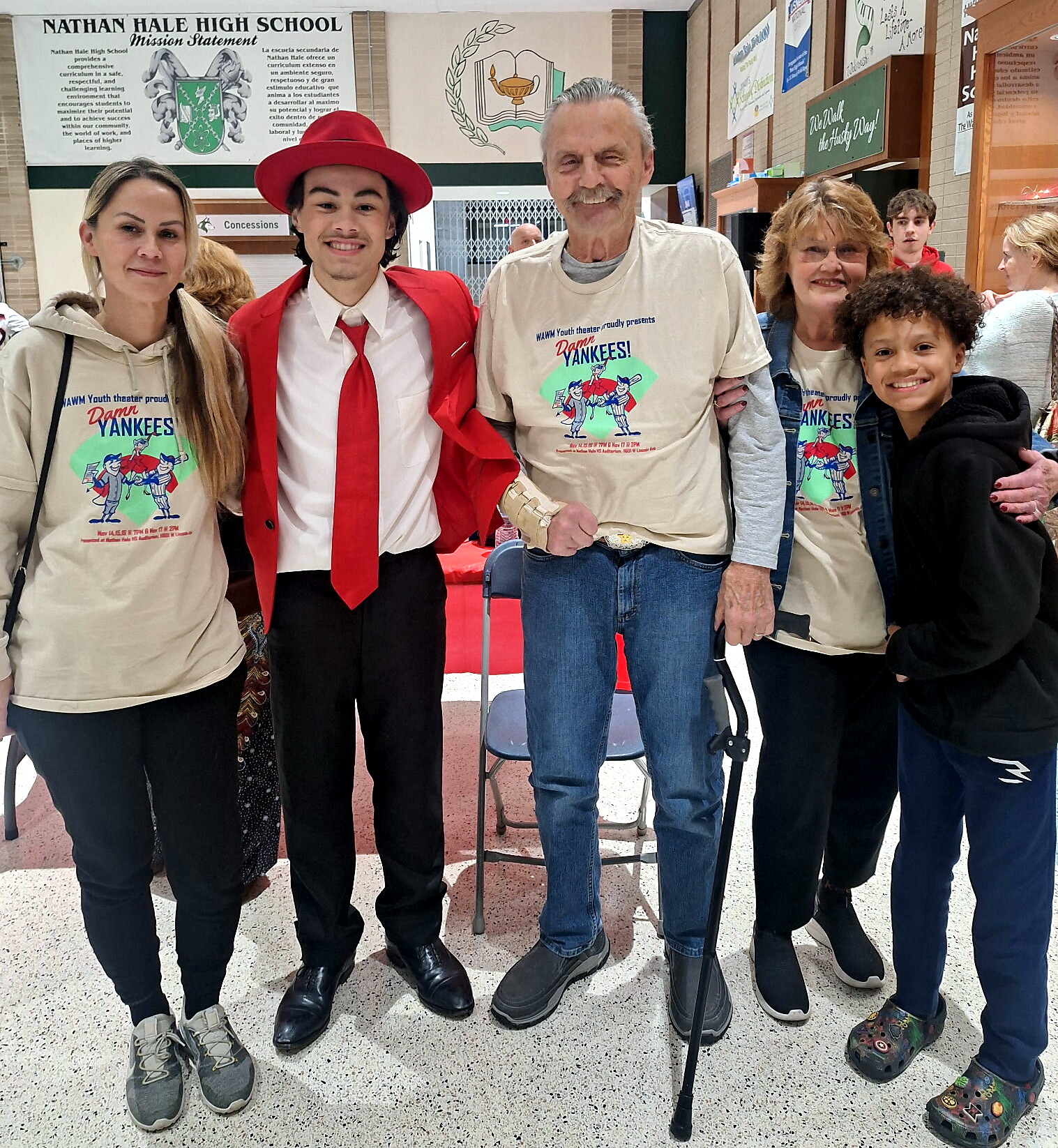 Five people posing in a school hallway; two wearing beige Damn Yankees shirts, one in a red suit and hat, and others smiling nearby with a crutch and brace visible in the center-right background.