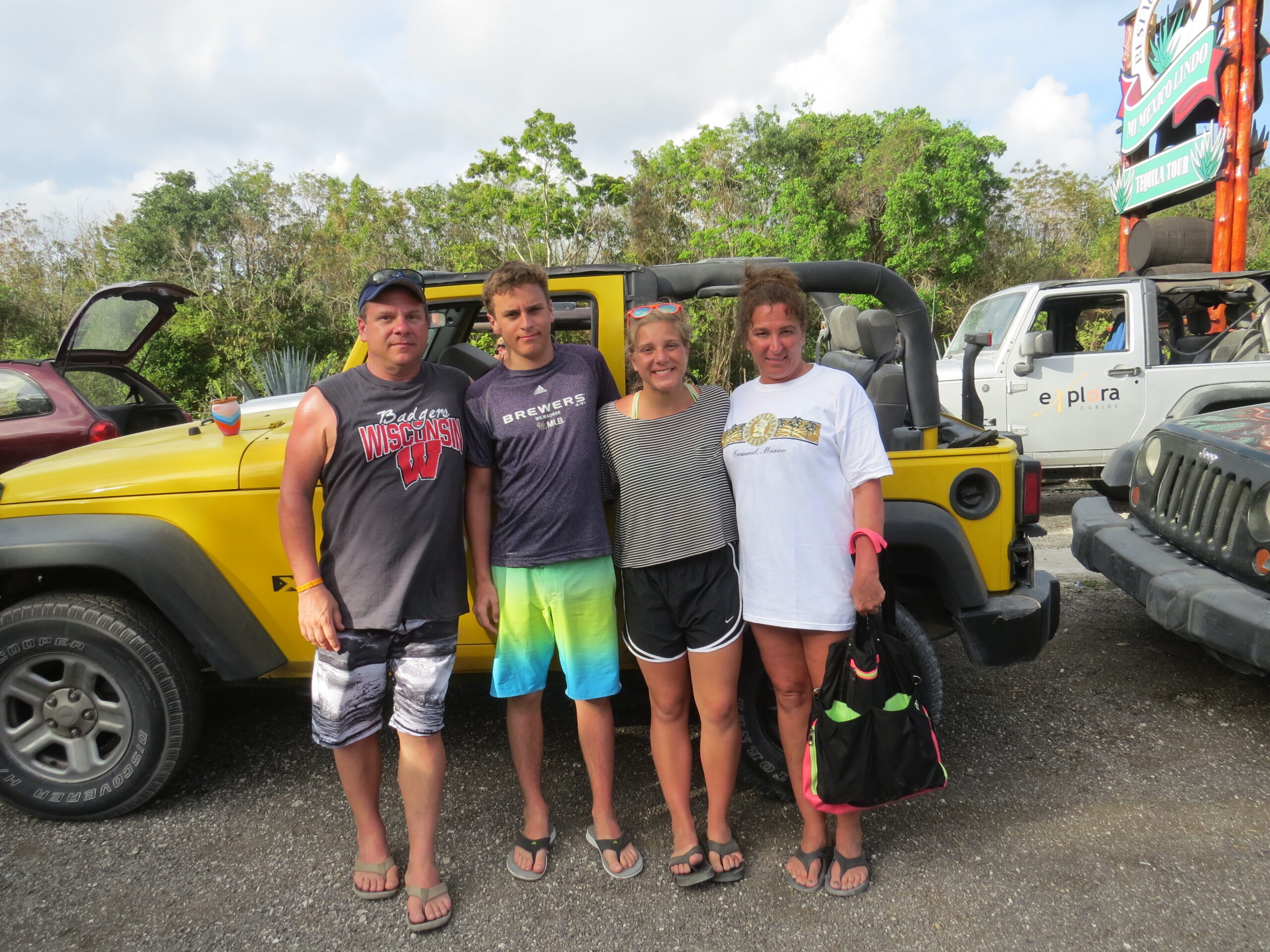 Four friends posing in front of a yellow jeep at an outdoor lot, smiling for the camera.