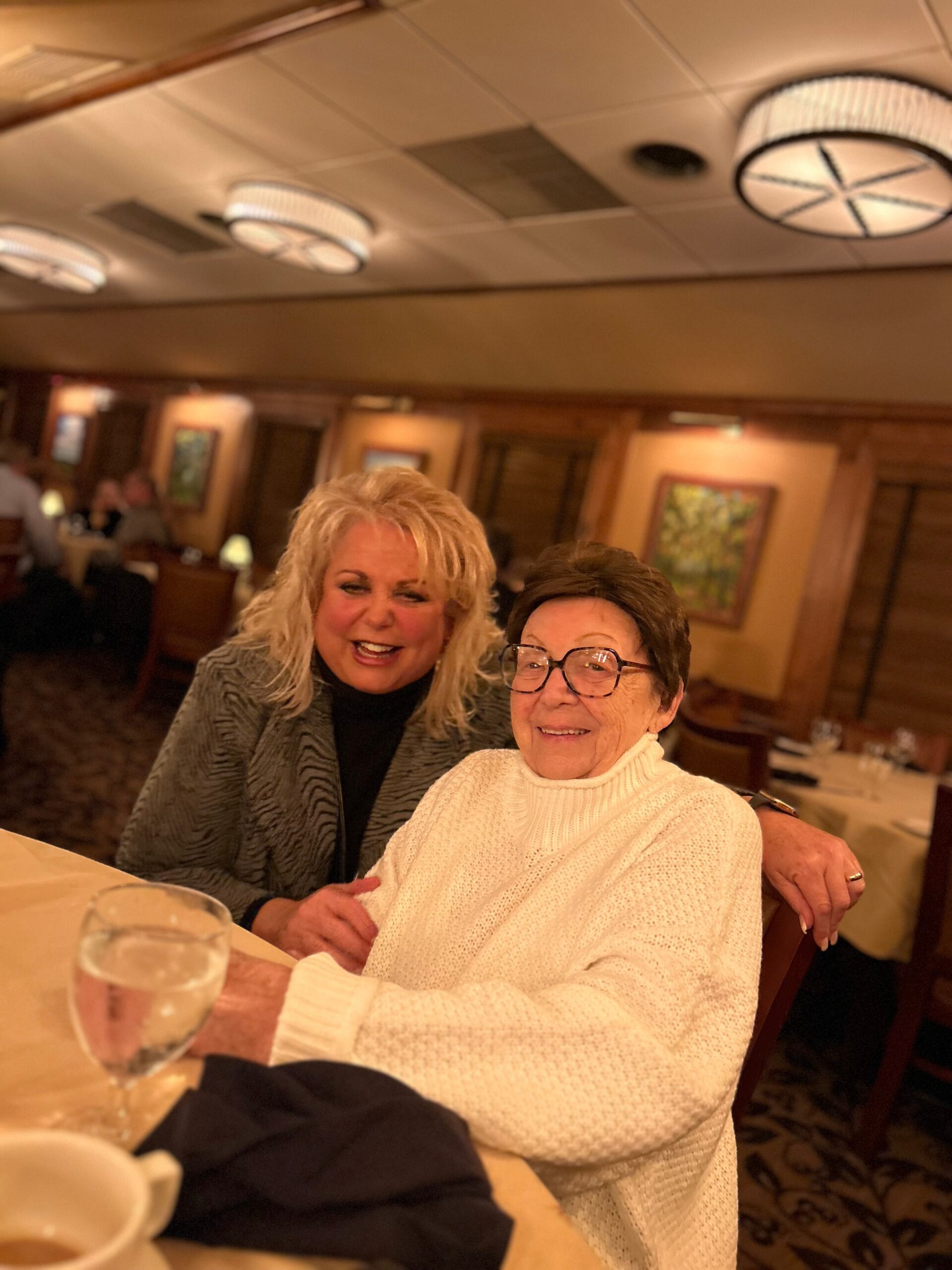 Two smiling women posing together at a restaurant table, with a glass of white wine in the foreground and warm, cozy lighting behind them.