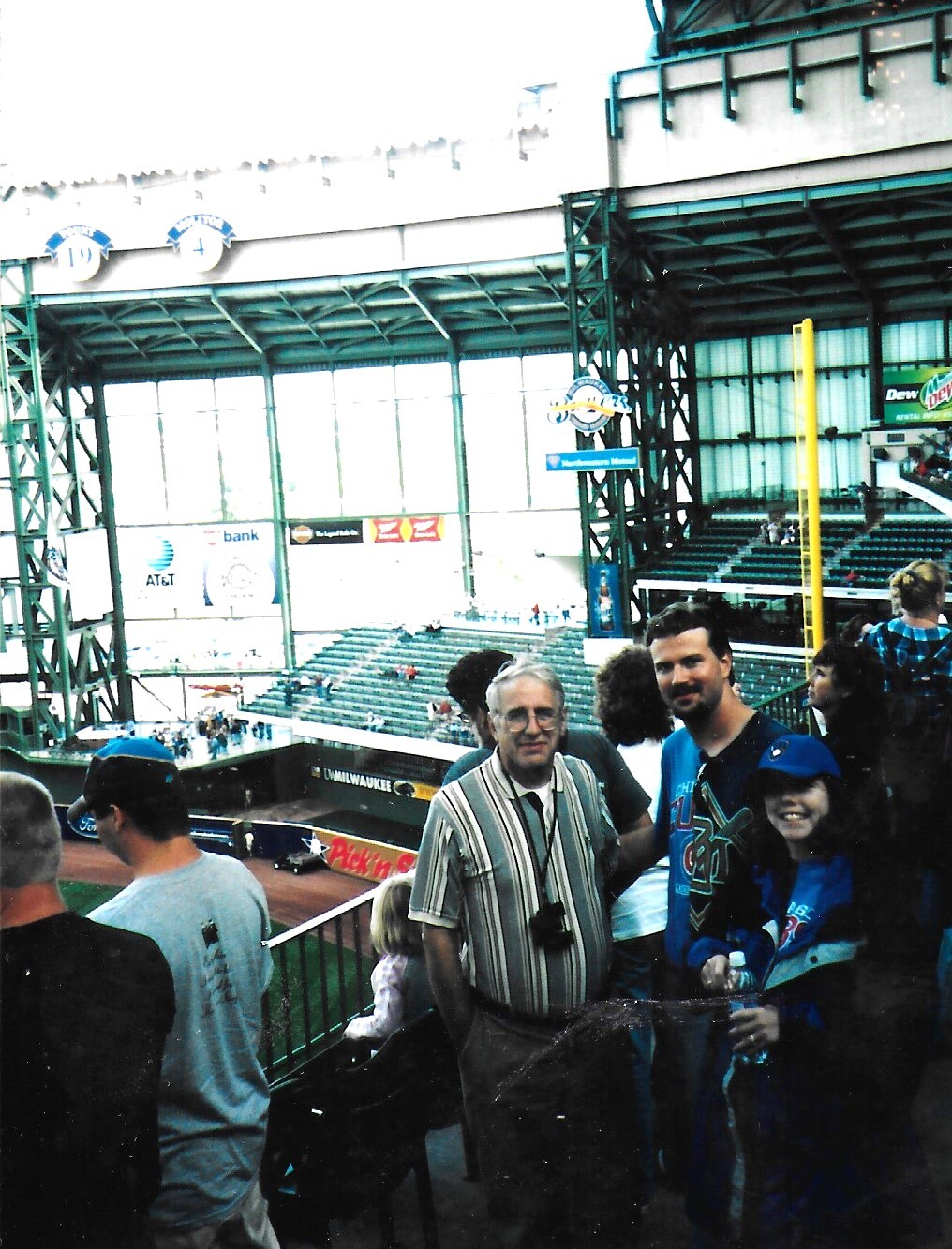 Group of fans posing for a photo at a baseball stadium with green seating and the field visible in the background.
