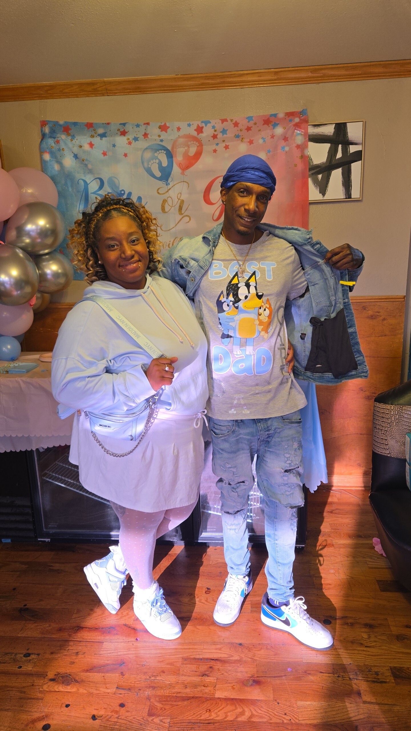 Smiling woman in a light hoodie stands next to a man in a denim jacket and blue durag at a party, with a 'Best Dad' graphic shirt and festive backdrop behind them.