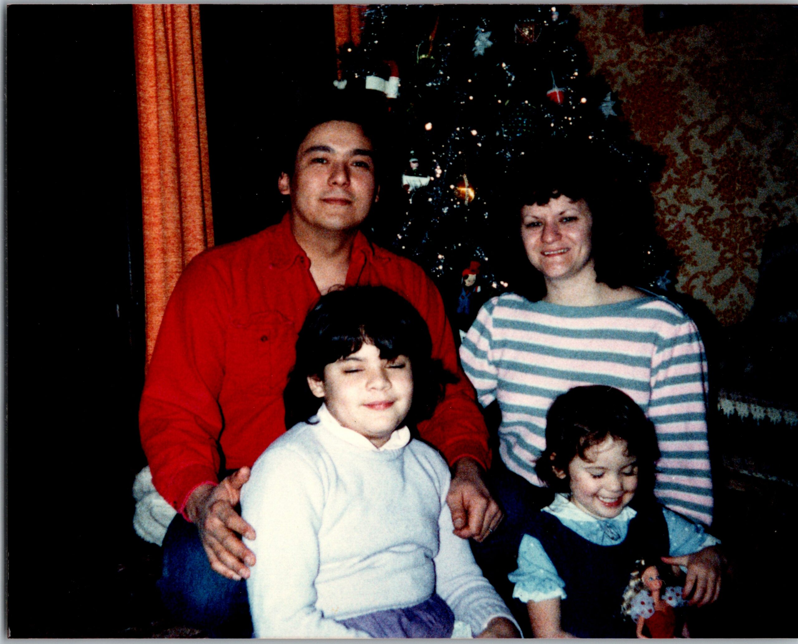 Family group photo indoors with a Christmas tree in the background; adults and children smiling together during the holidays.