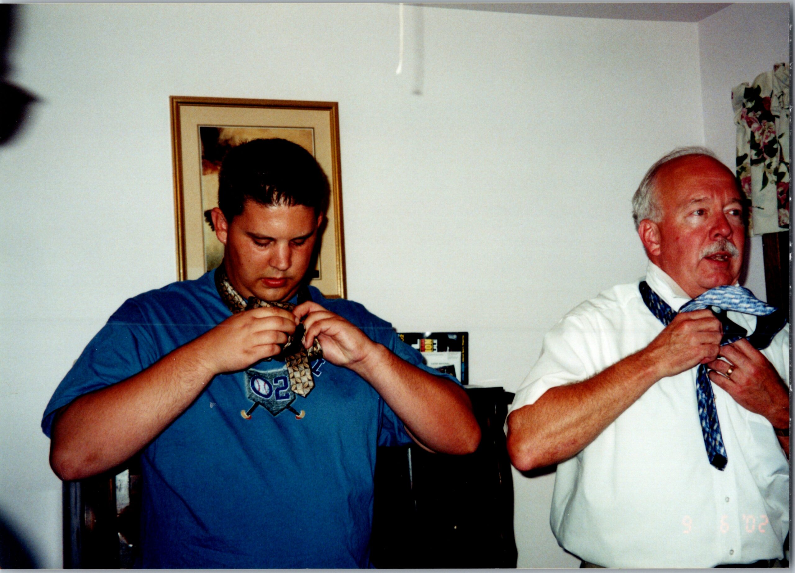 Two men in a room help each other adjust their ties; one in a blue shirt and one in a white shirt with a loosened tie, facing forward.