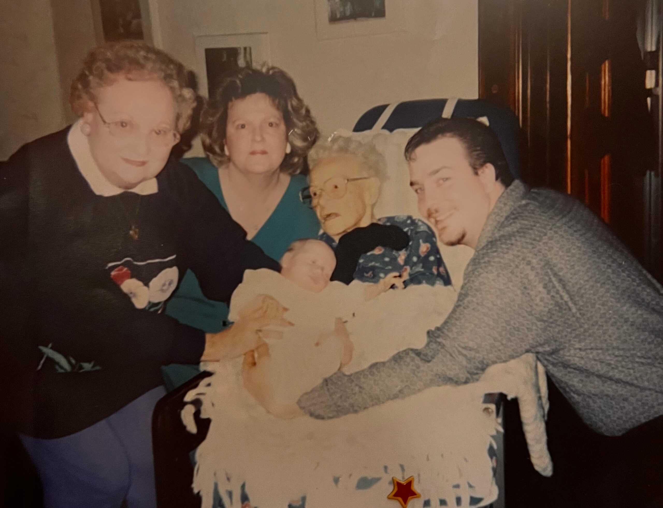 Family photo of three adults gathered on a couch with a baby, in a cozy living room setting.
