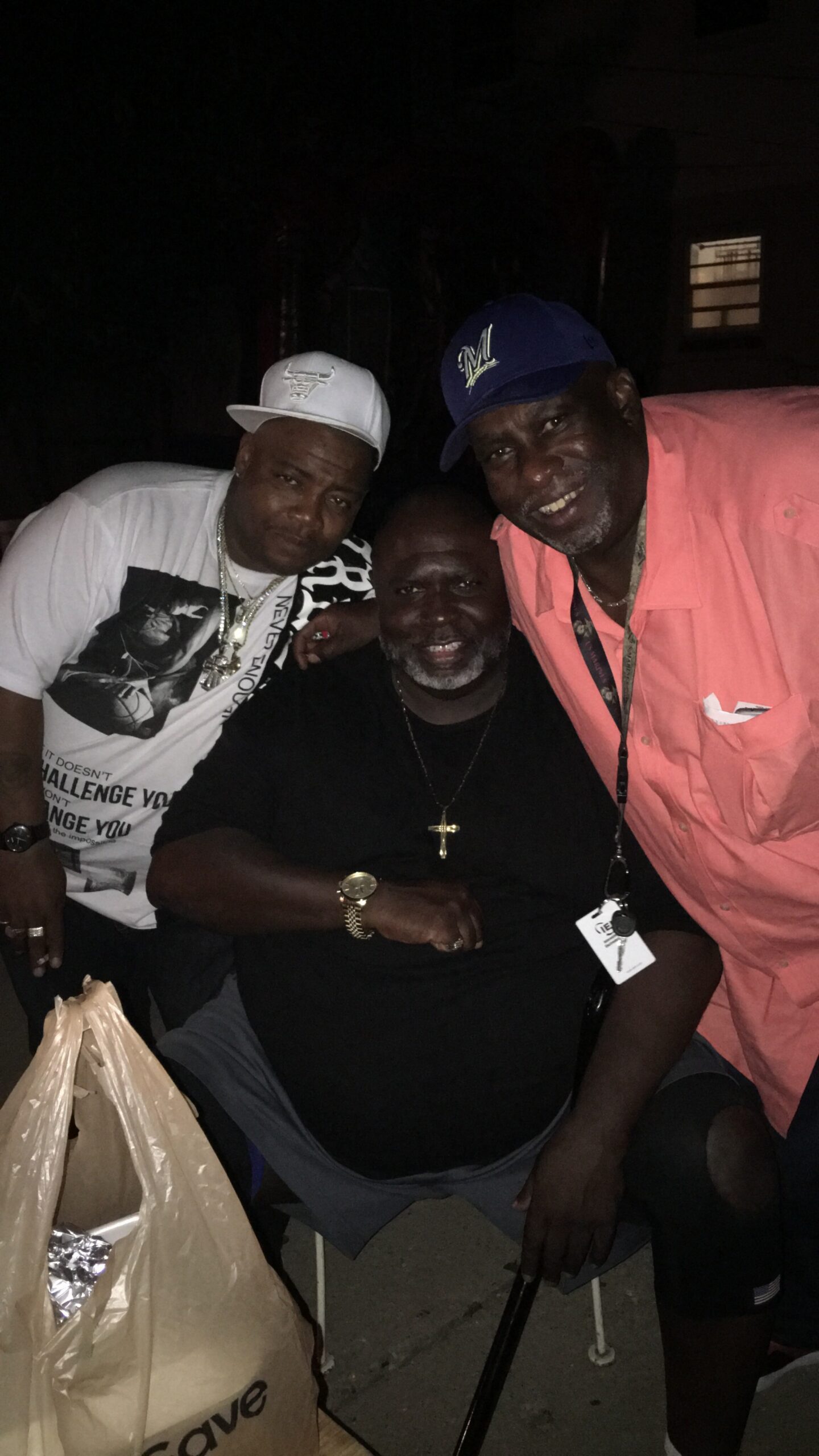 Three men posing together at night, two standing (white cap and blue Milwaukee cap) and one seated in black, all smiling beside a bag in the foreground, outdoors at night.