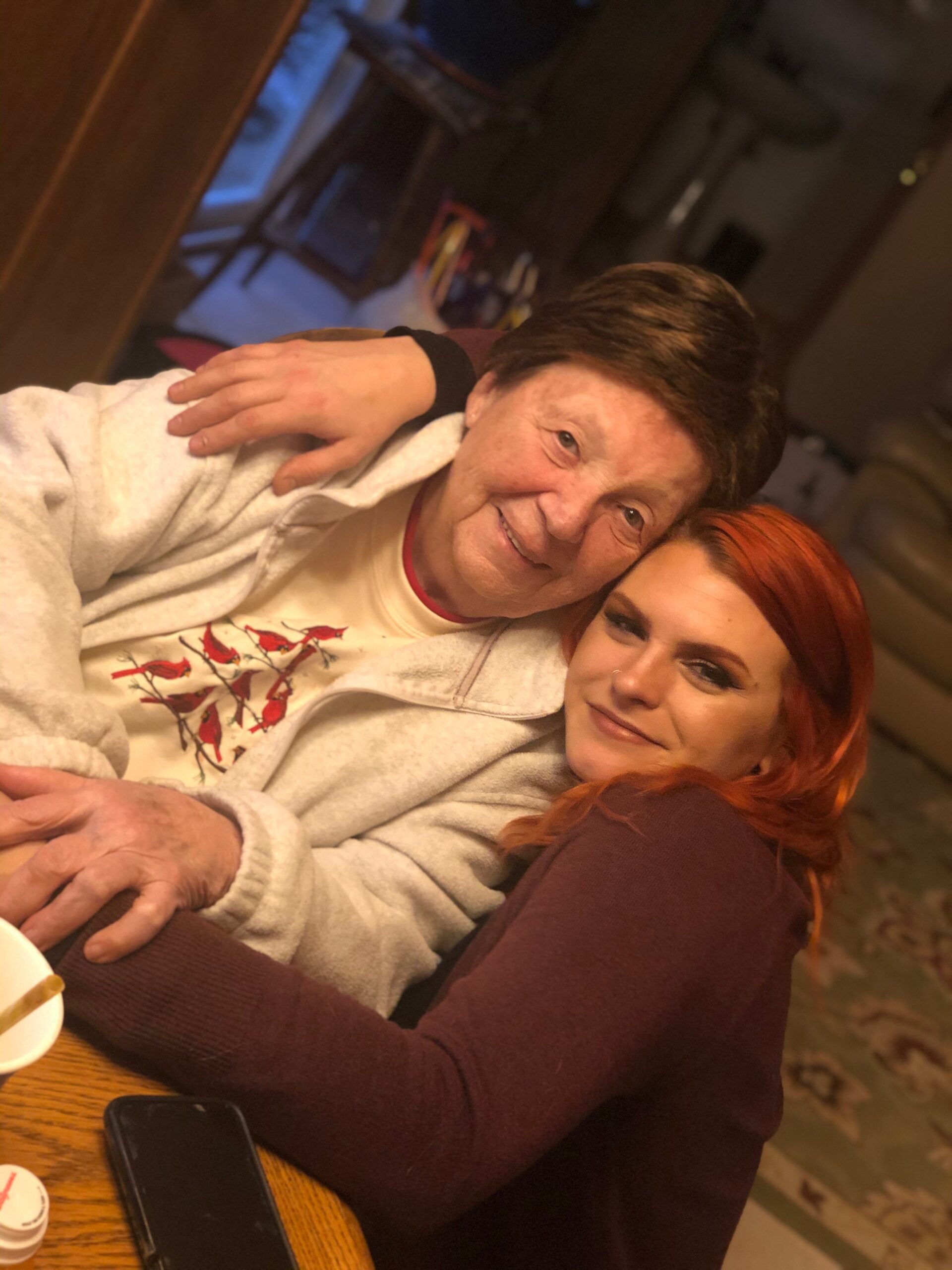 Two people cuddle and smile for a selfie at a table, a gray hoodie-wearing older man with a red bird design and a young woman with bright red hair leaning in together.