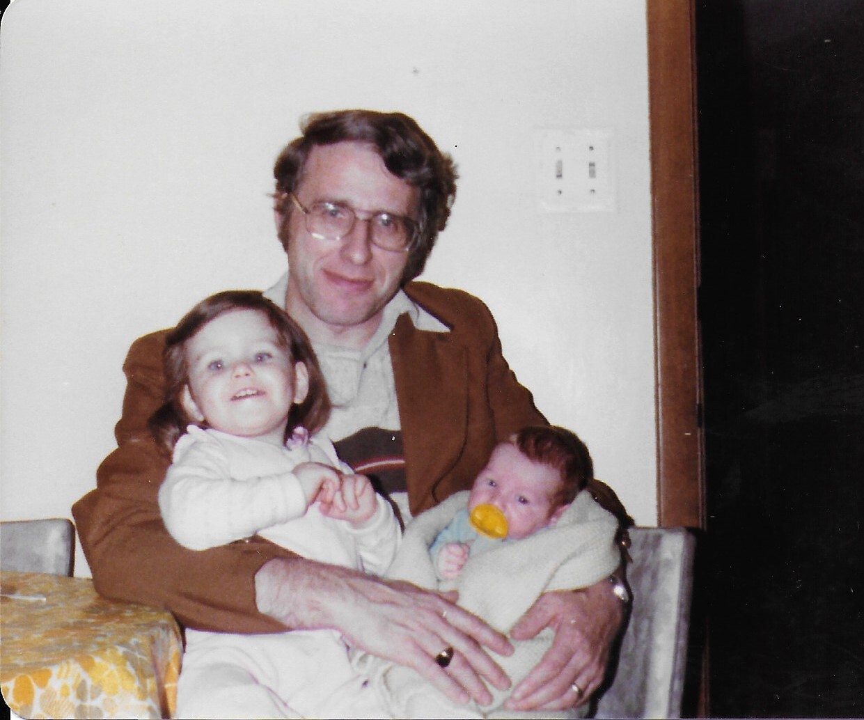 A man with glasses sits at a table, holding a young girl and a baby with a pacifier, in a cozy home setting.