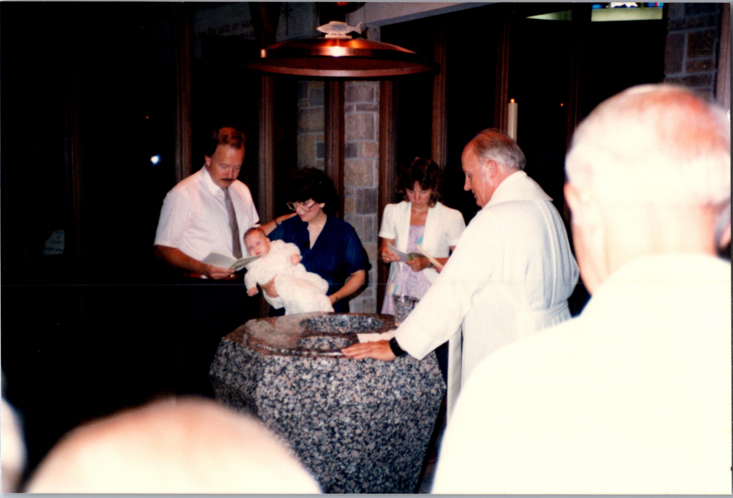 Family gathers for a baby baptism at a church; a woman holds the infant while others read from programs nearby.