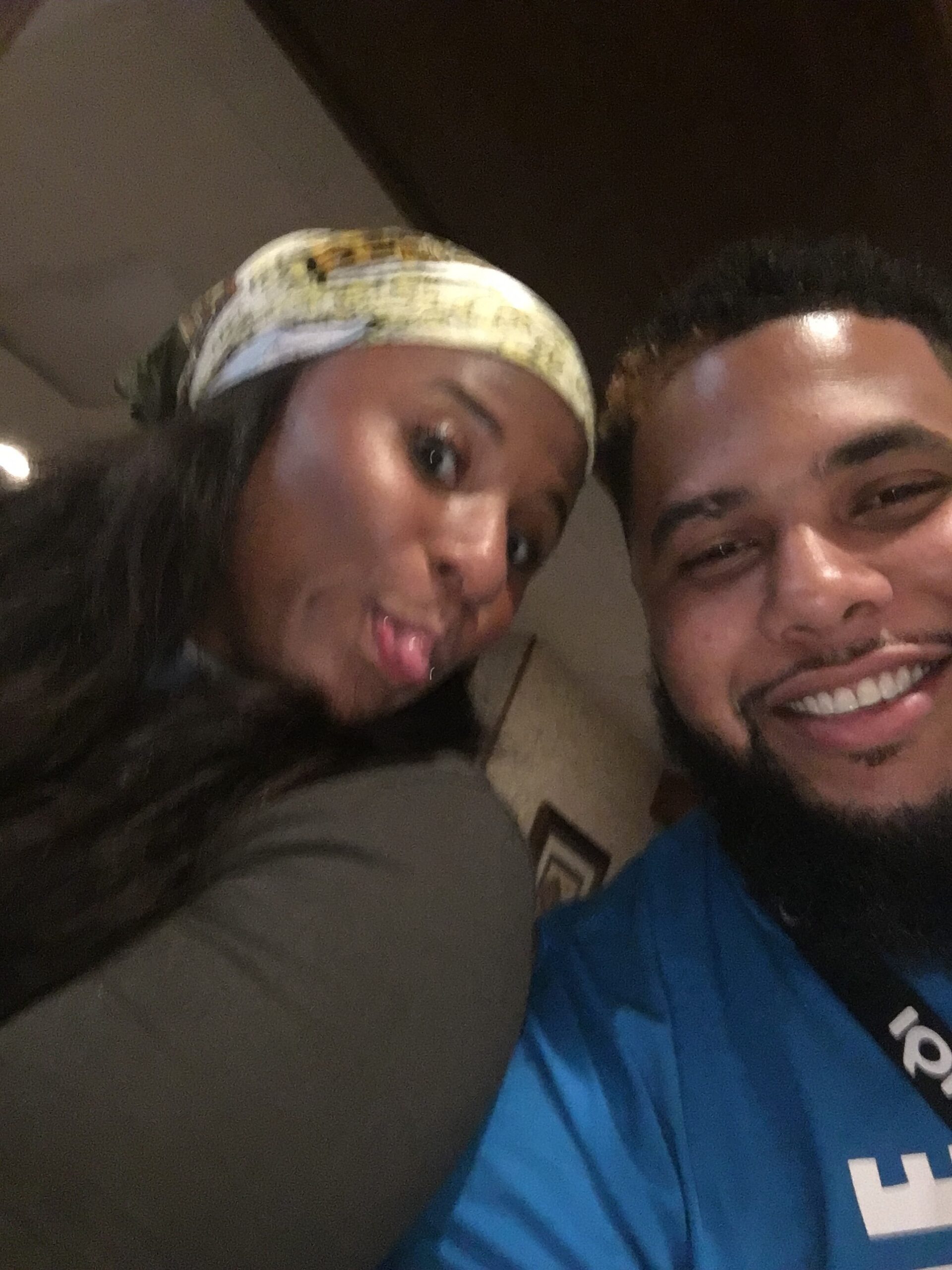 Close-up selfie of two friends, a woman with a patterned headband poking out her tongue and a smiling man with a beard in a blue shirt indoors.