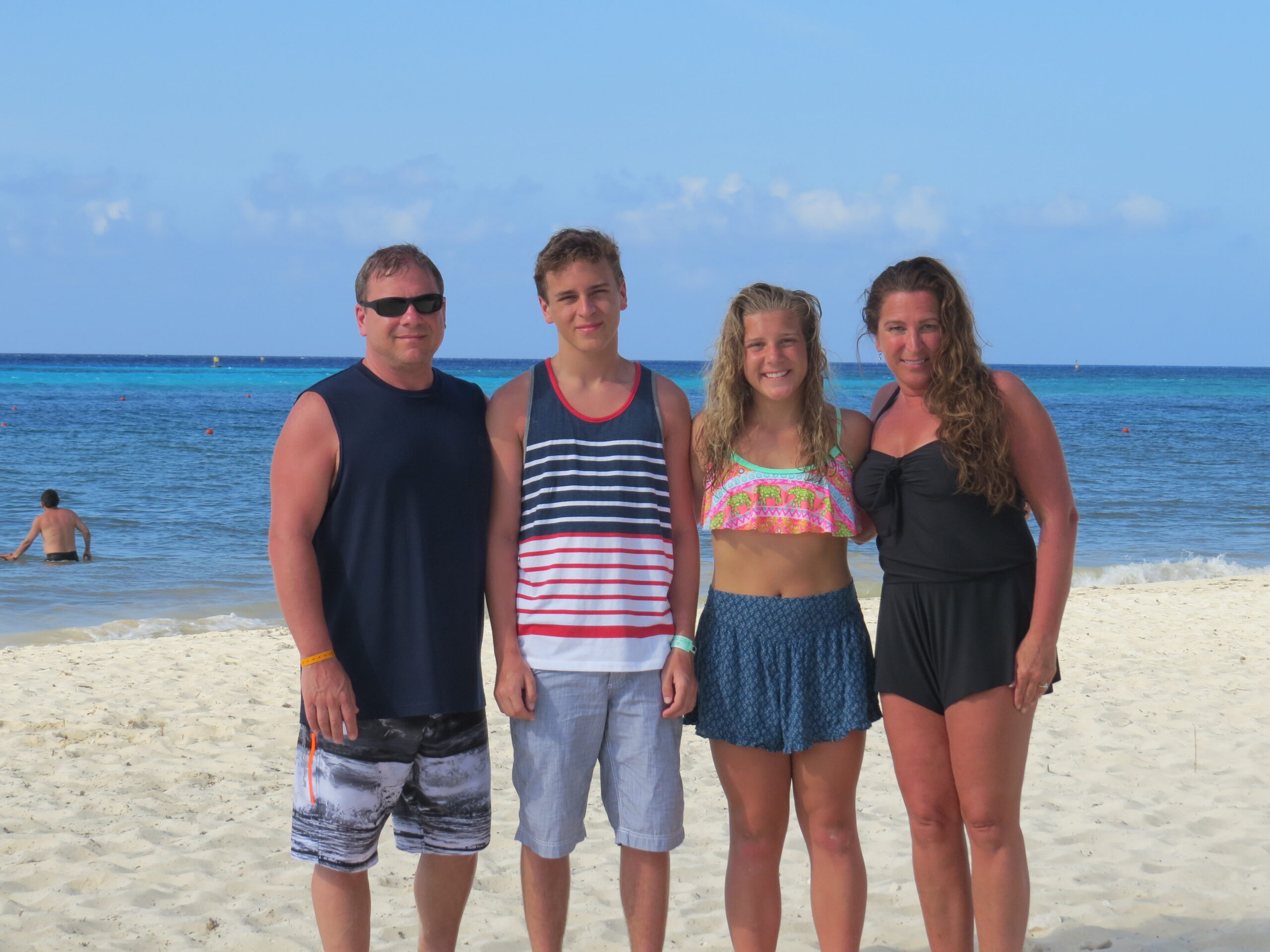 Four people stand side by side on a sandy beach with blue ocean water and a clear sky in the background. They wear swimsuits and summer clothing.