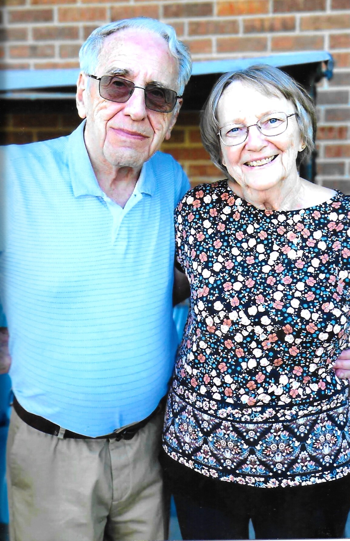 Smiling elderly couple standing side by side outdoors in front of a brick wall; man in a light blue polo, woman in a floral dress.