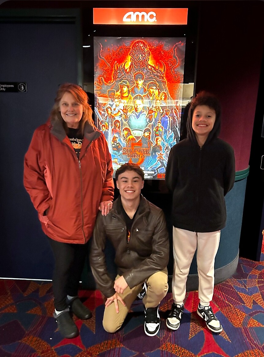 Three people pose and smile in front of a Stranger Things poster at an AMC cinema hall, with an orange AMC sign above them.