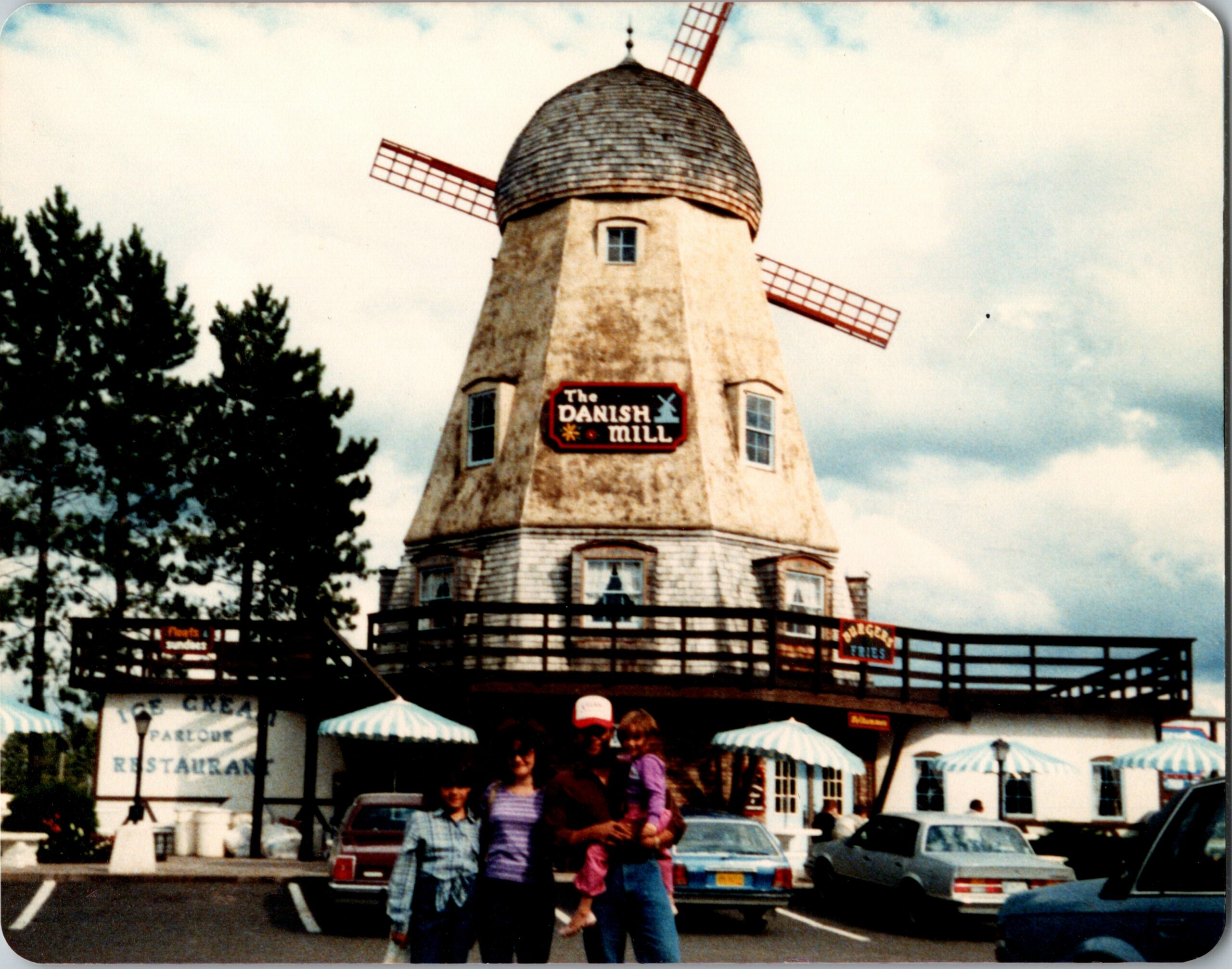 Exterior of The Danish Mill—a small windmill restaurant with a thatched roof and red sails, people posing in front under blue sky with trees nearby