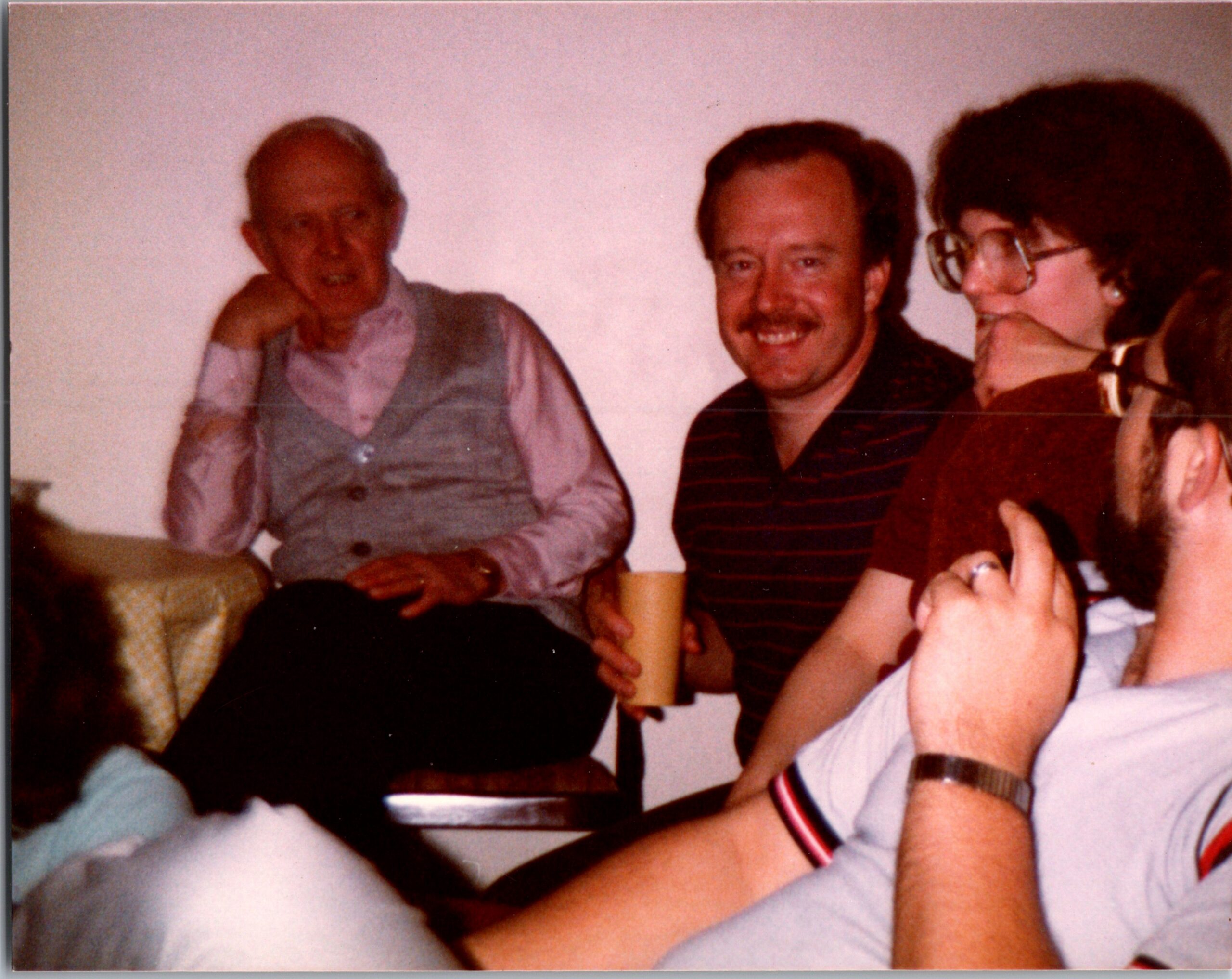 Three adults seated together at a casual gathering, the man in the middle smiling at the camera while others converse nearby.